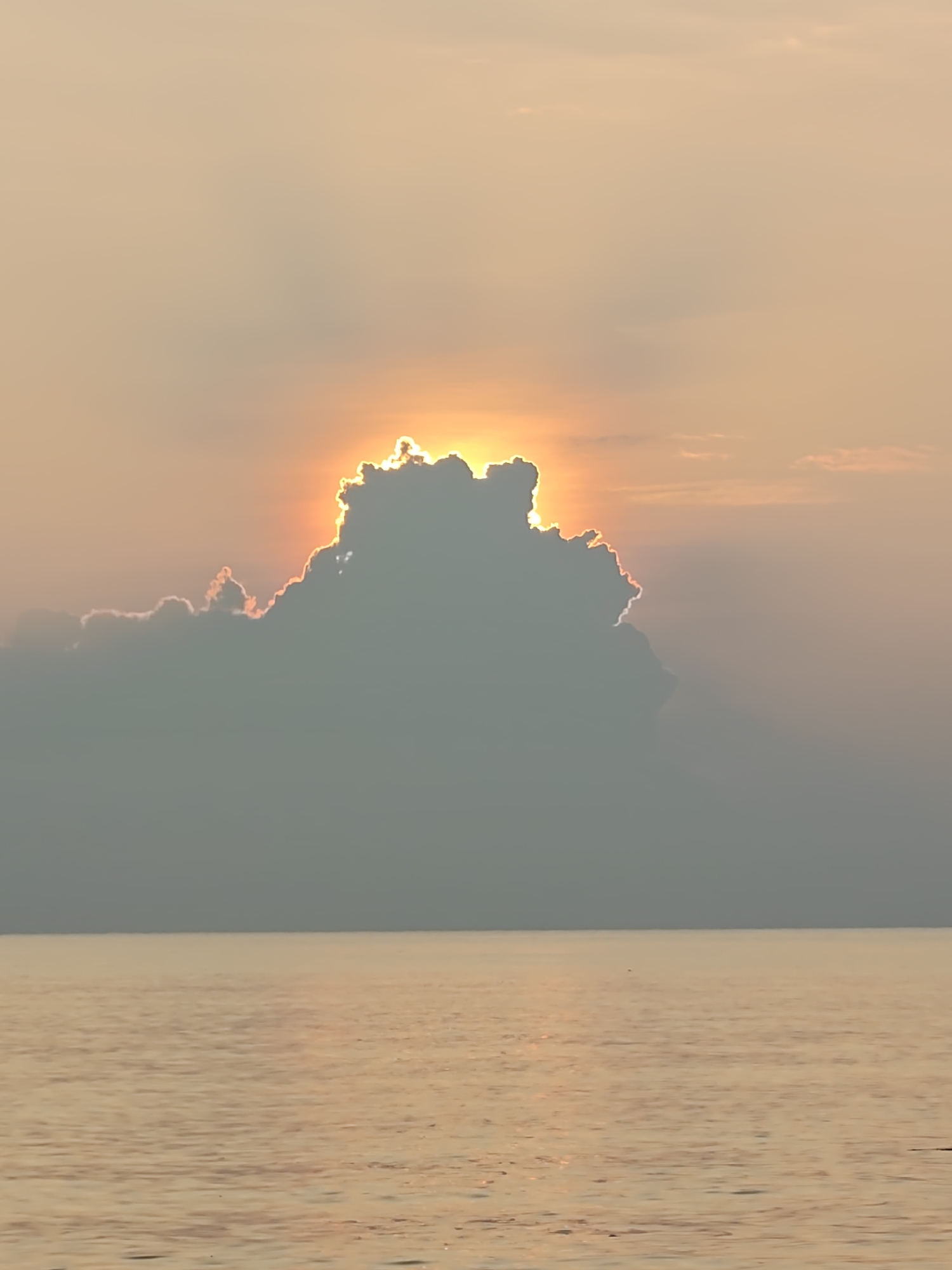 The image captures a sunset scene over the ocean, likely in Lhokseumawe, Indonesia. A large, dark cloud dominates the center of the frame, partially obscuring the sun, which shines brightly around its edges. The sky is a gradient of soft orange and peach hues, reflecting in the calm waters of the ocean. The horizon line is visible where the sea meets the sky, creating a serene and somewhat hazy atmosphere. There are no people or specific objects visible beyond the natural elements of clouds, sun, and sea.