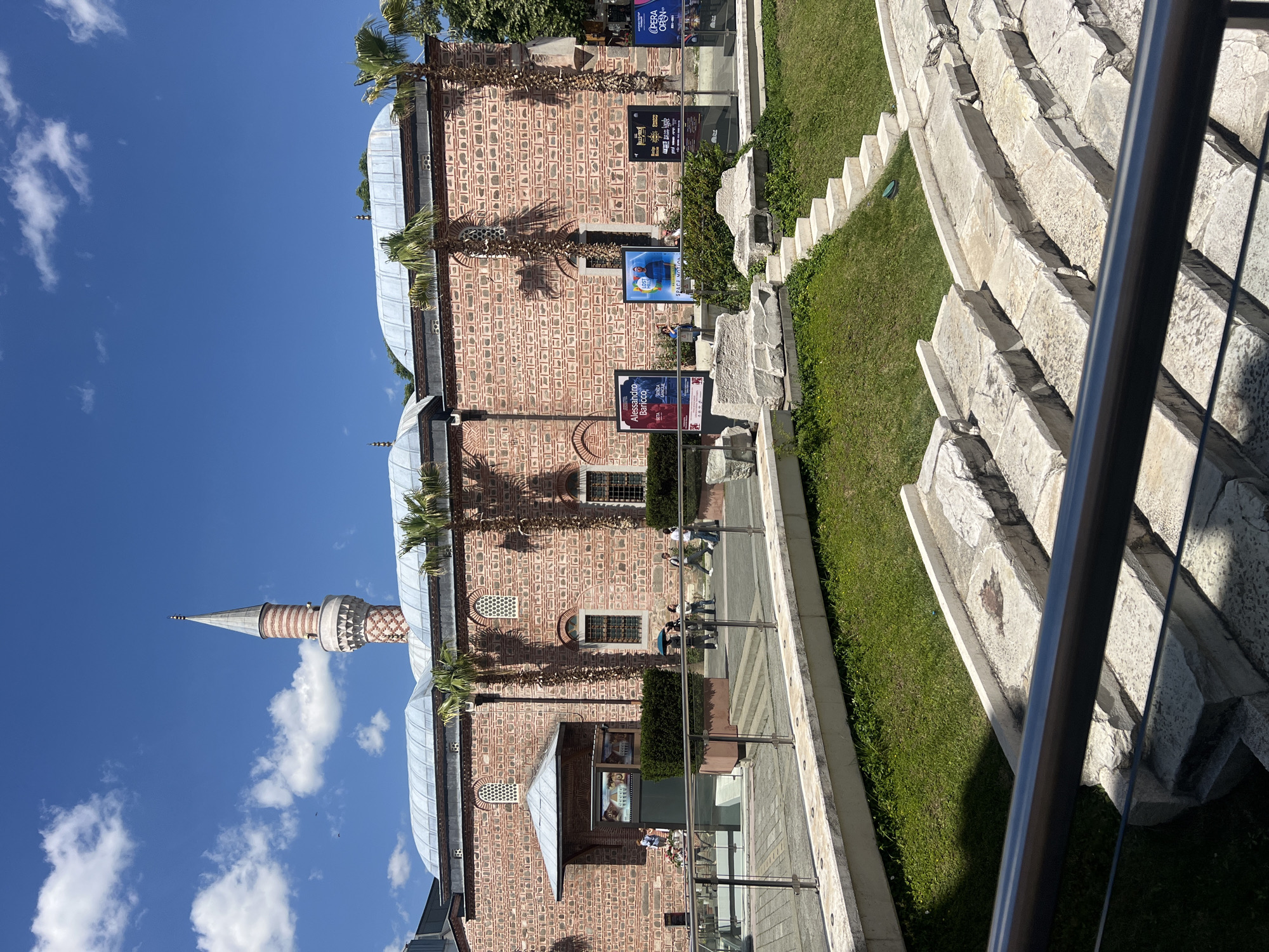 The image depicts the Dzhumaya Mosque in Plovdiv, Bulgaria, on a sunny day. The mosque's exterior is primarily red brick with decorative patterns. It features a minaret topped with a pointed spire, rising against a blue sky dotted with white clouds. Palm trees grow along the side of the mosque. Several posters and signs are affixed to the exterior walls. A stone structure with steps runs parallel to the mosque. Grass is planted in the spaces between the steps. A metal railing is visible in the foreground. People are walking on the pavement beside the mosque.