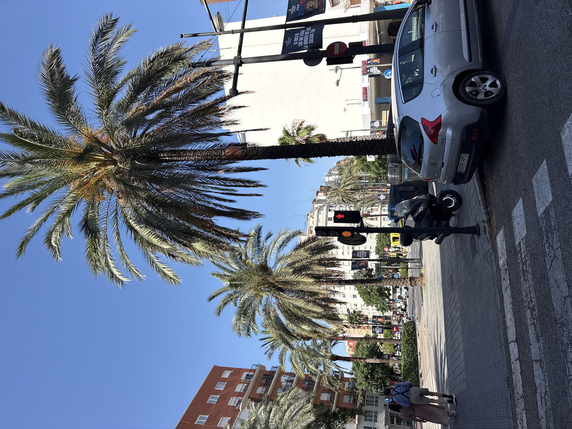 The image captures a street scene in Valencia, Spain, likely during daylight hours under a clear blue sky. Several palm trees are prominent, with their fronds reaching towards the sky. A silver Peugeot 3008 hatchback is parked on the right side of the road, facing left. A pedestrian crossing is visible in the foreground.
On the left side of the image, a multi-story brick building with balconies can be seen. Further down the street, more buildings and trees line the road. There's a traffic light displaying a red pedestrian signal, indicating that pedestrians should not cross. A dark-colored scooter is parked near the curb, adjacent to some trash receptacles. Two pedestrians, a man and a woman, are walking away from the camera on the sidewalk. In the background, traffic and other pedestrians are visible, suggesting an active urban environment. Banners are displayed on poles, some with text and graphics, typical of city advertising. The overall impression is a sunny, urban street typical of a Mediterranean climate.