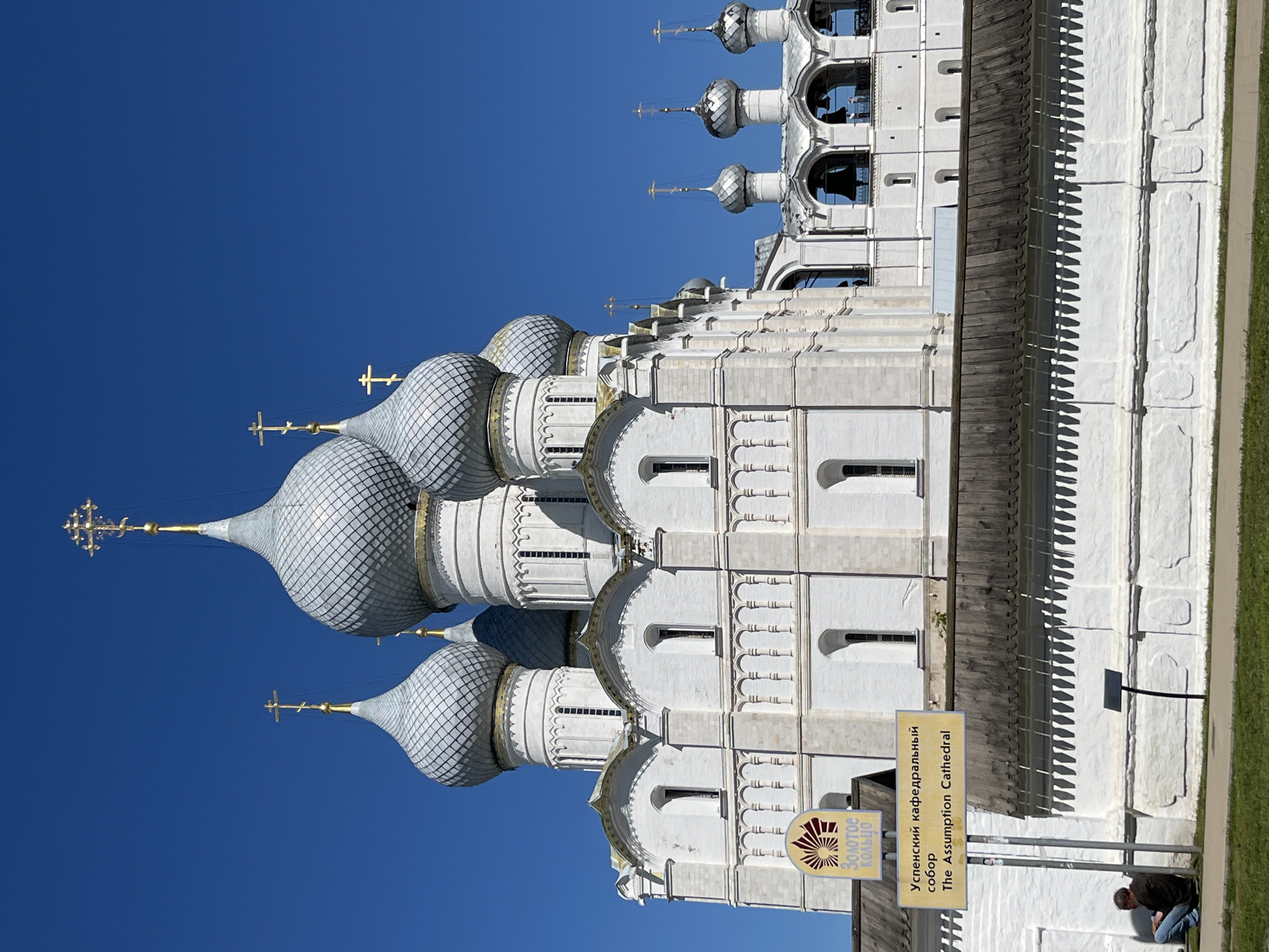 In Ново-Никольское, Russia, the Assumption Cathedral is a grand white cathedral with multiple silver domes topped with golden crosses. The cathedral is surrounded by a white brick wall and a wooden fence. A sign in front of the cathedral reads "Успенский кафедральный собор The Assumption Cathedral" in Russian and English. A man is sitting on the ground near the sign, leaning against the wall. The sky is clear blue.
