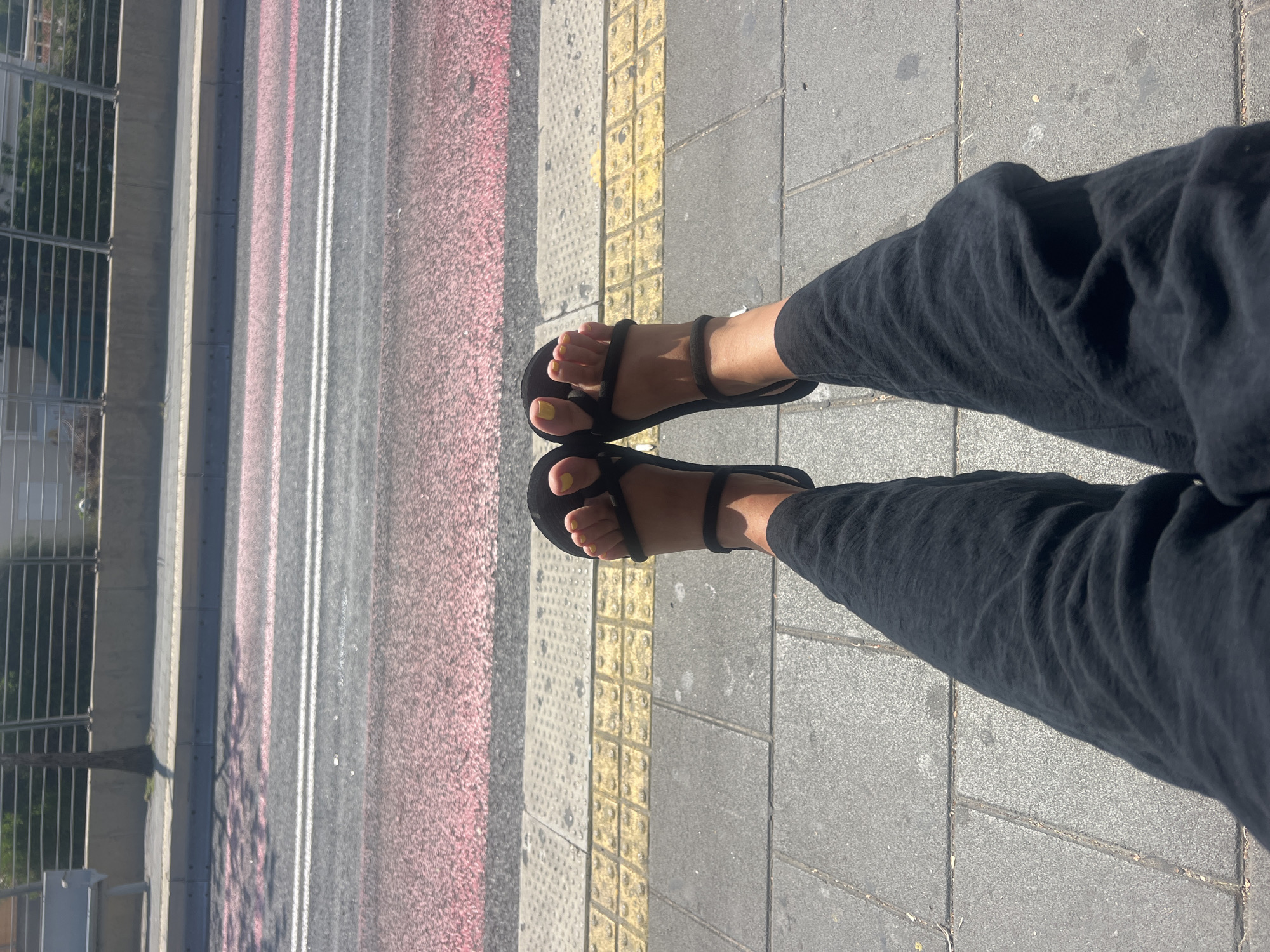 A person is sitting on a paved sidewalk in Kiryat Motzkin, Israel, with their legs extended towards a street with a red and white painted surface. The person is wearing black pants and black sandals, and their toenails are painted yellow. The sidewalk is made of gray concrete pavers with tactile paving inserts. In the background, there is a street with painted markings and a metal fence. The scene appears to be an outdoor public space.