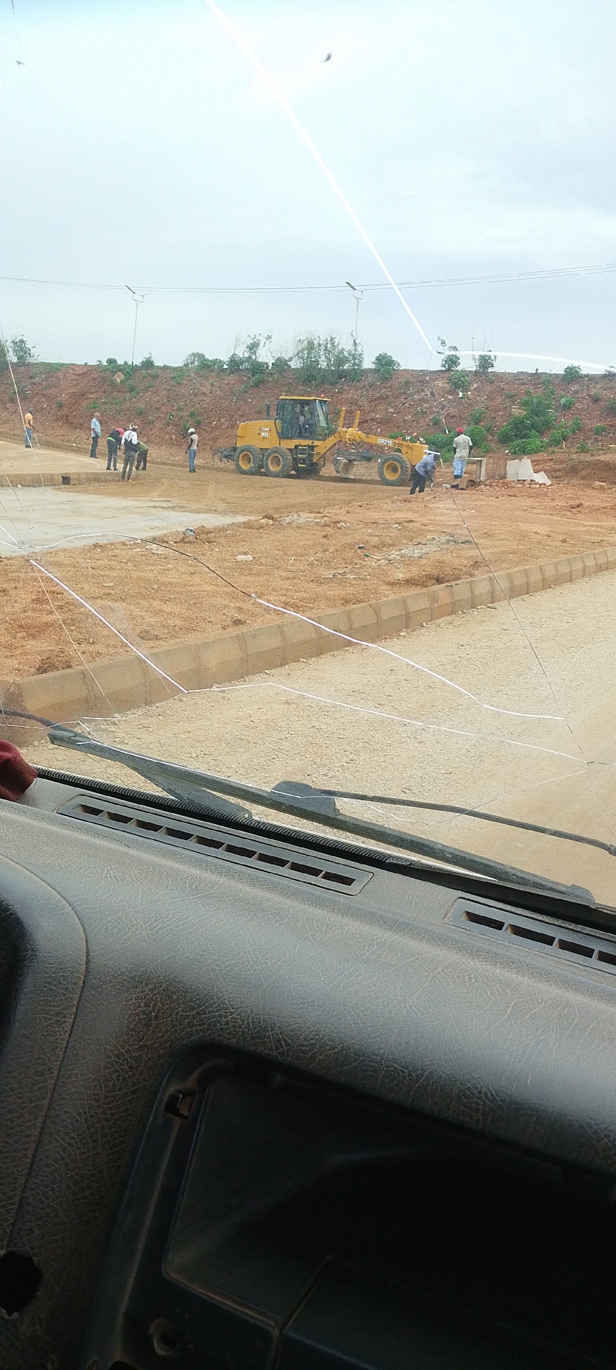 An exterior, eye-level, medium shot captures a construction site under a hazy, overcast sky. In the middle ground, a yellow grader is actively leveling a dirt surface, with several construction workers standing nearby. The foreground is dominated by the interior of a vehicle, showing a cracked windshield and part of the dashboard. The scene is set on a dusty, unfinished road, with a dirt embankment and some vegetation in the background.