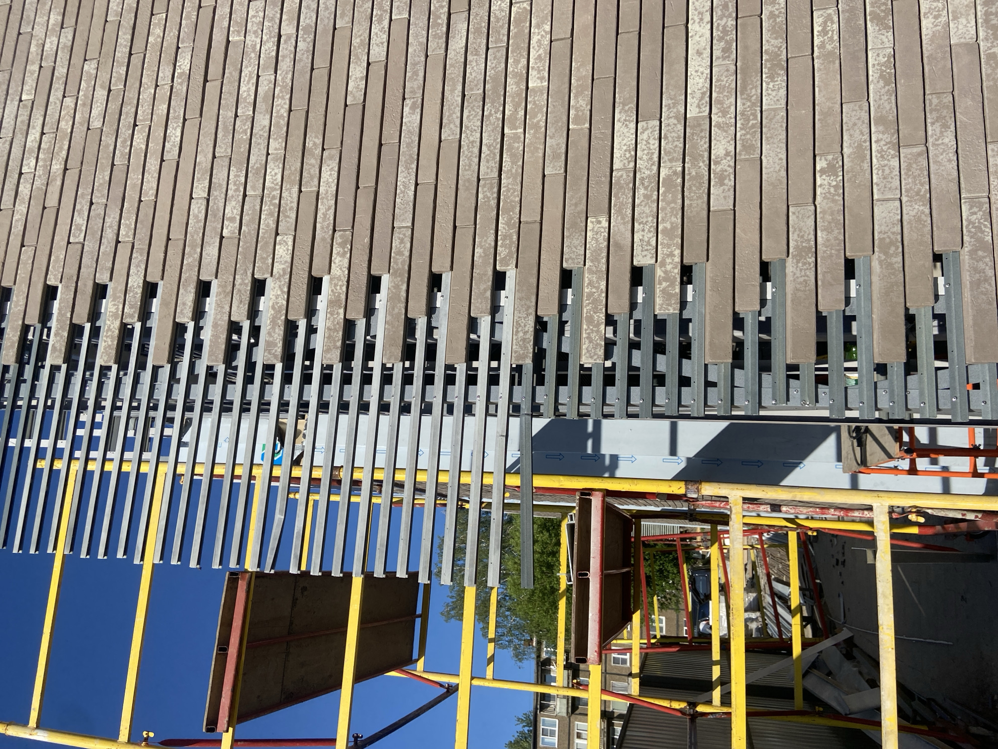 The image shows a close-up view of a building facade under construction. The facade is made of vertically arranged, rectangular, tan-colored bricks. Below the bricks, there's a series of vertical, gray, metal slats or bars. Scaffolding, consisting of yellow and red poles and crossbars, is visible in the lower portion of the image, along with some boards or panels attached to the scaffolding. Through gaps in the construction, a glimpse of a blue sky is seen, along with trees and buildings that appear to be part of an urban environment. The image is rotated. There is no information to confirm the location is in Kazan, Russia.