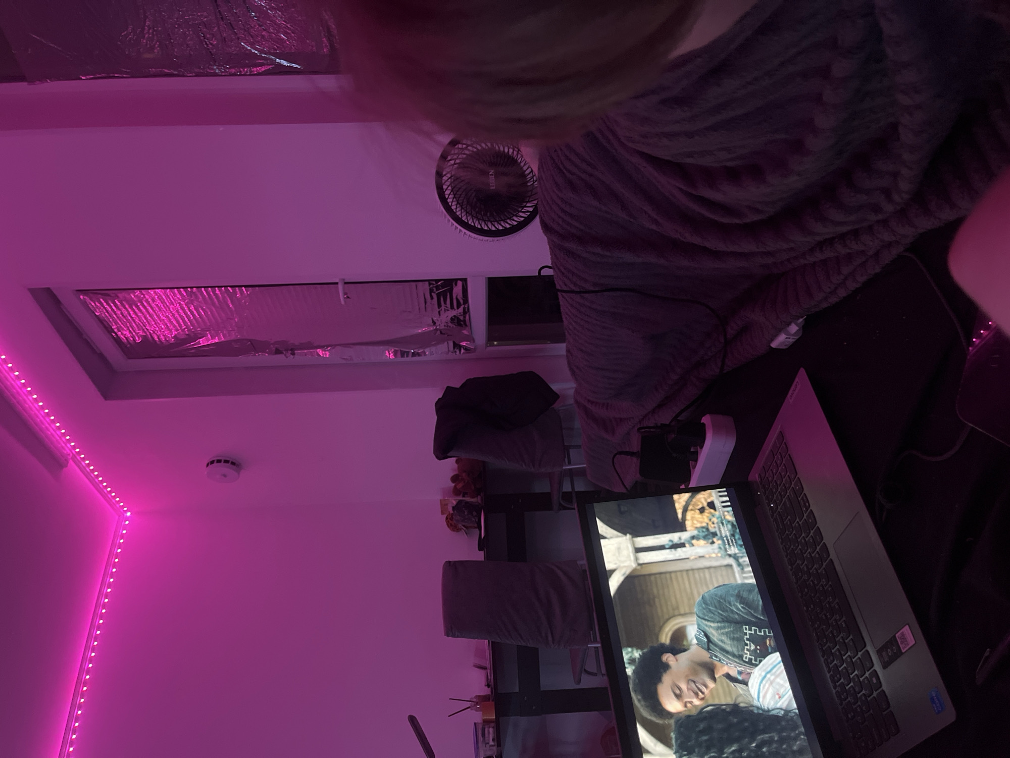 A room with pink lighting. The ceiling has foil-covered panels, a fan, and a smoke detector. A laptop displaying a man is on a desk. Two chairs are present. A person covered in a blanket is partially visible in the upper right. No specific location in Saint Petersburg, Russia, is identifiable.