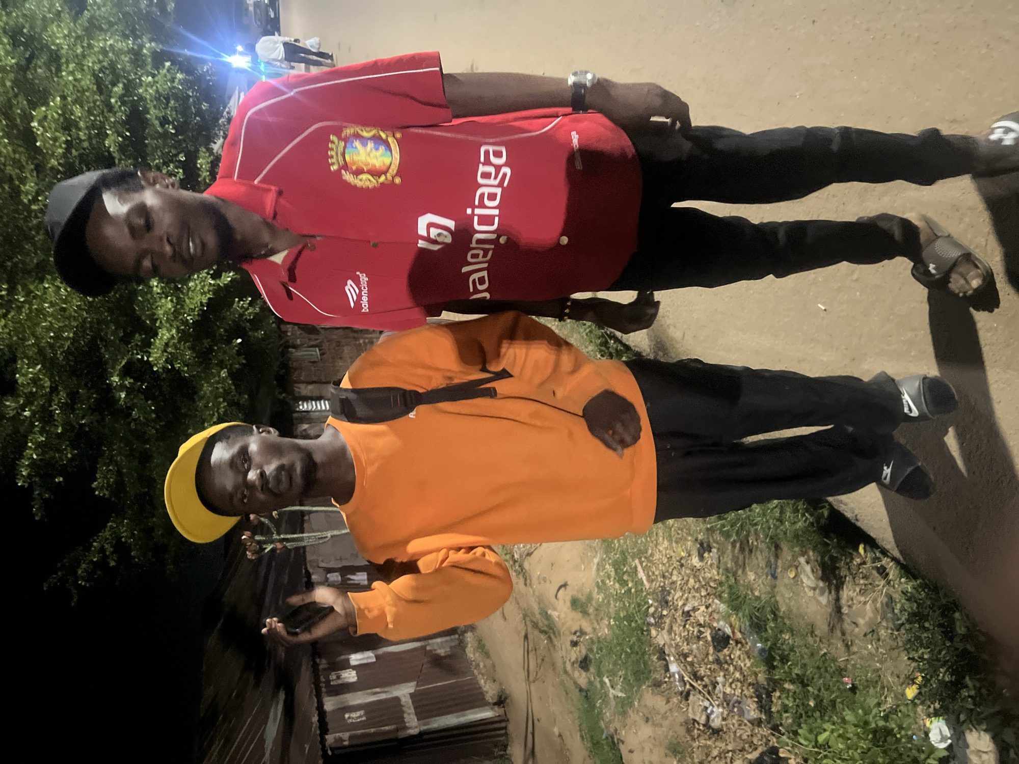 The image displays two young men standing outdoors at night in Gura Top, Nigeria. The scene is illuminated by artificial light sources, casting bright glares, and features a ground composed of dirt, sparse grass, and scattered litter. Corrugated metal structures and green foliage are visible in the background.
The man on the left wears a red short-sleeved polo shirt, featuring a prominent crest and the word "Balenciaga" written vertically on its side. He is also wearing black trousers, a black baseball cap, and grey slides with an "Under Armour" logo. A watch is visible on his left wrist. He has a beard and a neutral expression, looking slightly downwards.
The man on the right is dressed in an orange long-sleeved sweatshirt, black trousers, and black slides with a "Nike" swoosh logo. He wears a black baseball cap with a visible yellow brim and has a black fanny pack or sling bag slung across his chest. He holds a smartphone in his right hand and looks towards the camera with a neutral expression.
Both men appear to be casually posing for a photograph in the nighttime setting.