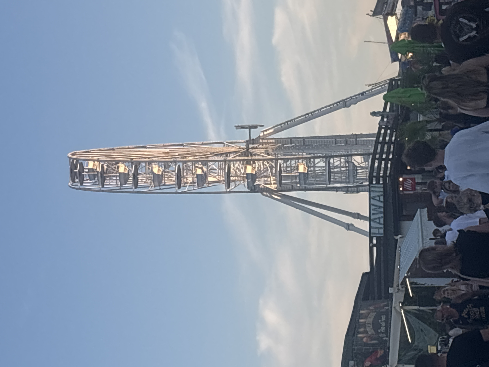 The image shows a Ferris wheel at what appears to be a fairground or amusement park in Nesebar, Bulgaria. The Ferris wheel is tall and white, with several gondolas visible. In the foreground, there are several people walking around. Some of the people are looking at the Ferris wheel, while others are looking at their phones. There are also some food stalls and stands set up. The sky is blue with some clouds. The overall atmosphere is one of a lively summer day at an outdoor event.