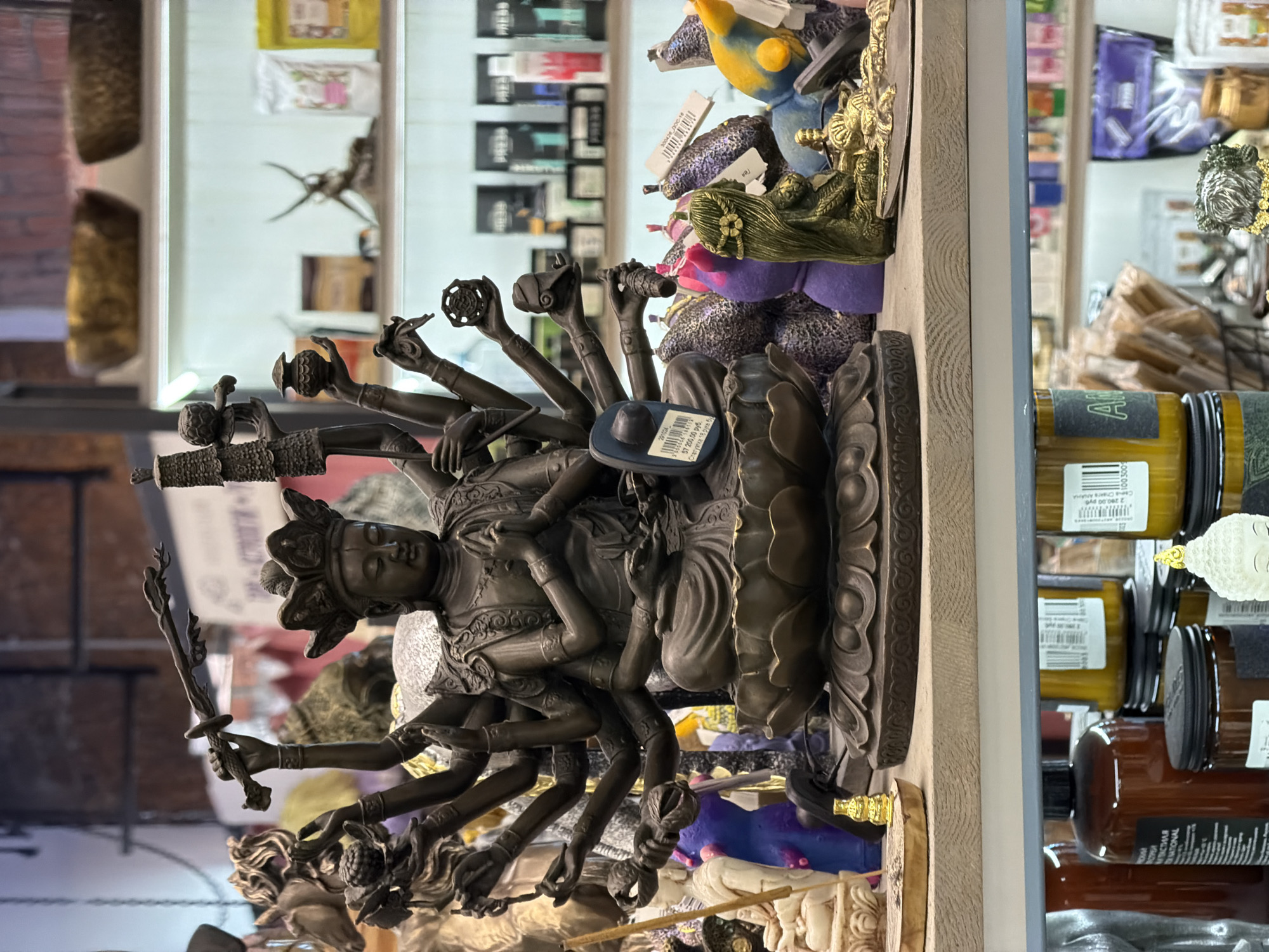 A bronze statue of a multi-armed deity, possibly a representation of Shiva or another Hindu god, is displayed in what appears to be a shop window or showcase in Moscow, Russia. The statue is surrounded by various other items, including food products like jars of honey or jam. The OCR text indicates the item's price in rubles. The statue has a complex design with several arms holding different objects and is seated on a lotus base. The background is a mix of other merchandise and reflections in the glass of the showcase.