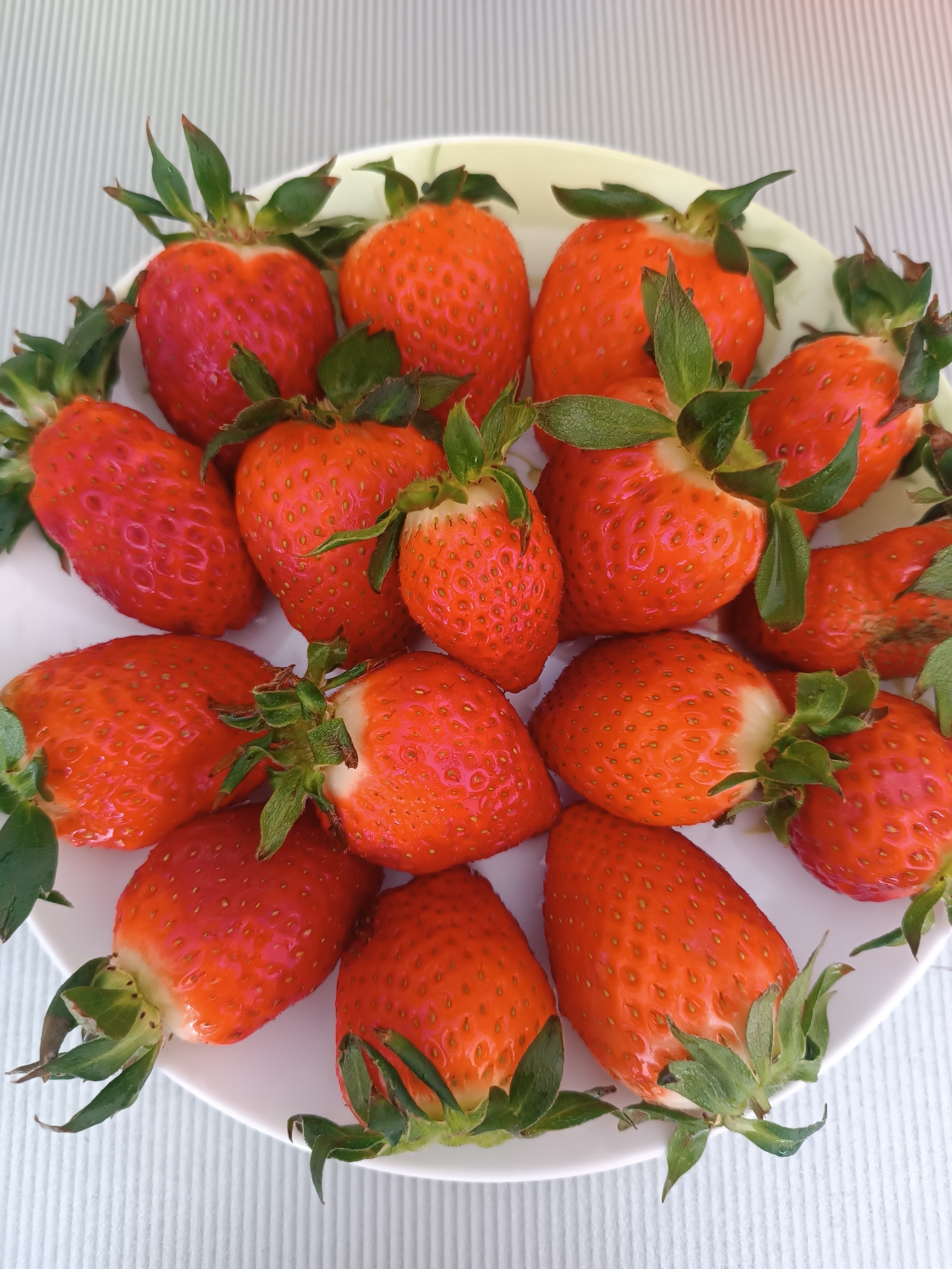 The image captures a white, round plate filled with approximately fifteen to twenty ripe strawberries, located in the city of Chaikovsky, Russia. The strawberries are predominantly vibrant red, with various shades including darker crimson and lighter orange-red tones. Each berry retains its green calyx (leafy cap) and a short portion of the stem. Some strawberries exhibit a slightly paler, yellowish-white area at their top where they connect to the calyx. Small, yellow achenes are visible across the surface of the fruit. The plate rests on a light-colored, vertically striped or ridged surface, likely a tablecloth or placemat, under soft, ambient illumination. No individuals are present in the scene, and no specific actions are being performed.