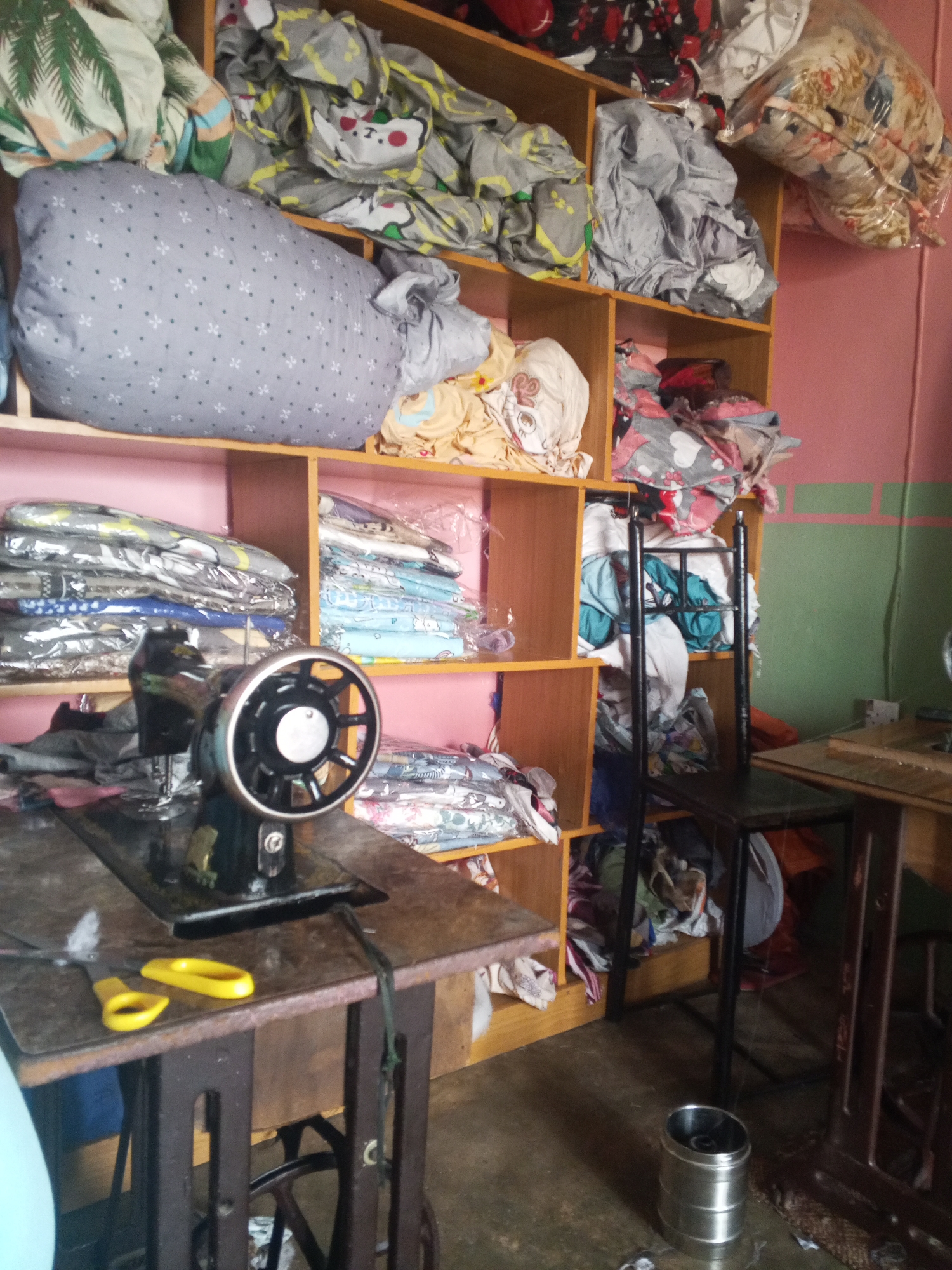 The image depicts a room filled with shelves stacked with various fabrics and bedding. In the foreground, a vintage sewing machine sits on a wooden table, accompanied by a pair of yellow-handled scissors. A chair is positioned near the shelves. The environment appears to be a storage area or workspace, likely a tailor's shop or a home sewing room, given the presence of the sewing machine and abundant fabric. The walls are painted pink and green, with some shelves showing a wooden finish. The floor is made of concrete. There are no people visible in the image.