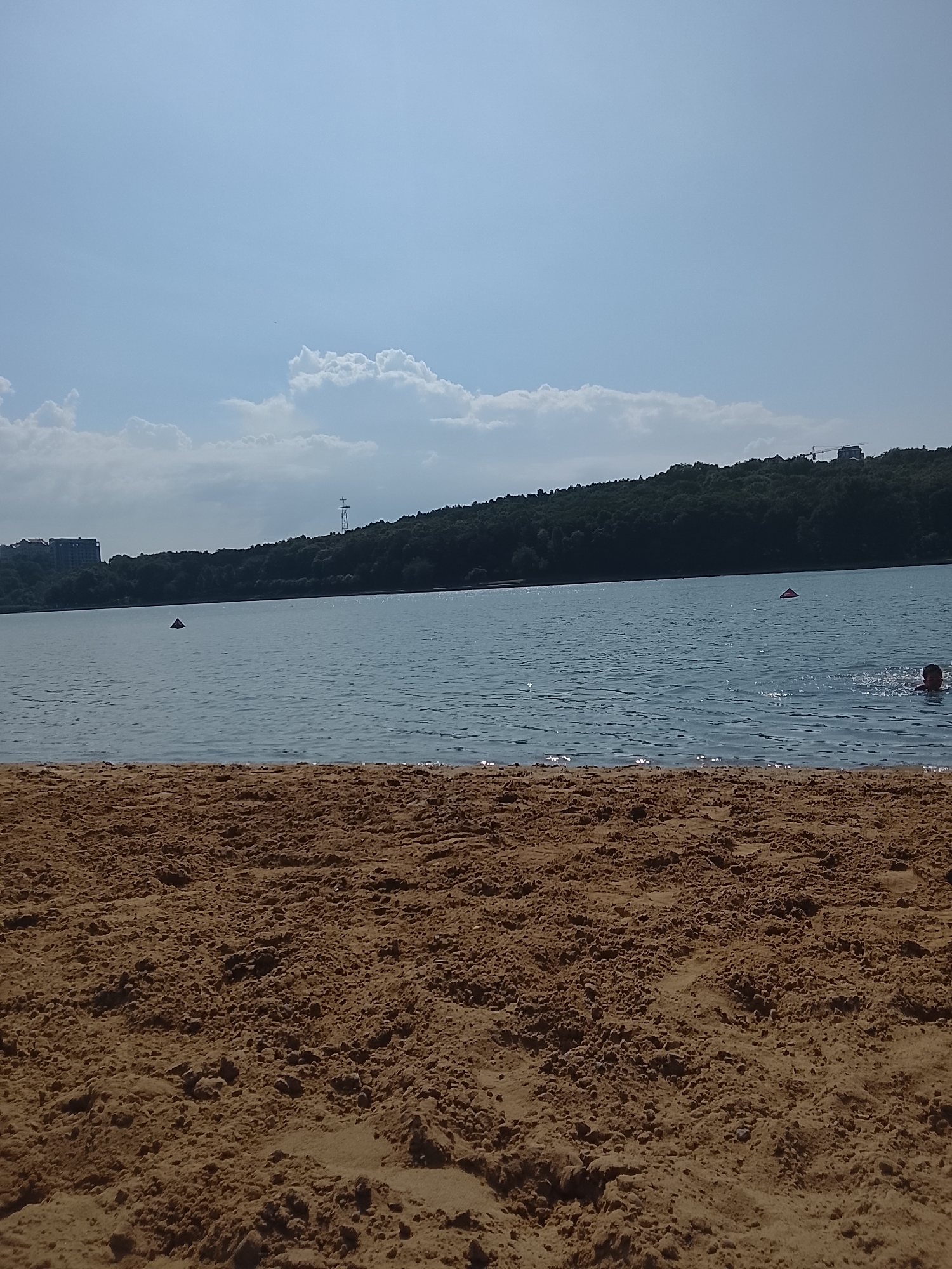 The image depicts a sandy beach area in the foreground, leading to a large body of water. In the distance, a densely wooded hill forms the backdrop, with some buildings visible on the left and right edges of the hill. The water is calm, with a few buoys or markers visible. On the right side of the water, a person is swimming, with their head and shoulders above the surface. The sky above is blue with scattered white clouds. The scene is likely a recreational area, possibly a lake or reservoir within or near the city of Chișinău, Moldova, given the presence of both natural and urban elements.
