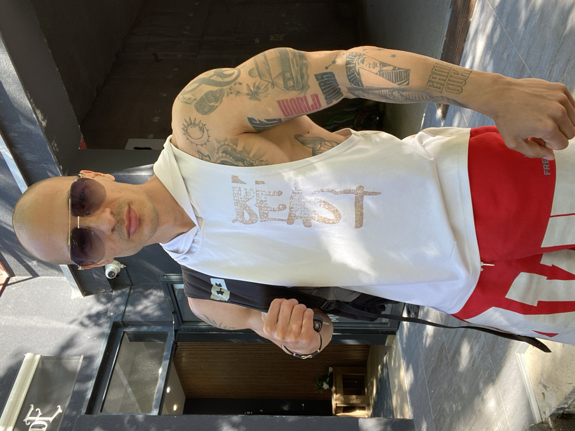 A muscular man with a shaved head, tattoos on his arms, and sunglasses stands in front of a building. He is wearing a white tank top with the word "BEAST" printed on it, along with red and white pants. He has a black backpack slung over his shoulder. The location is a modern building with a black facade and large windows, likely in the city of Pleven, Bulgaria.
