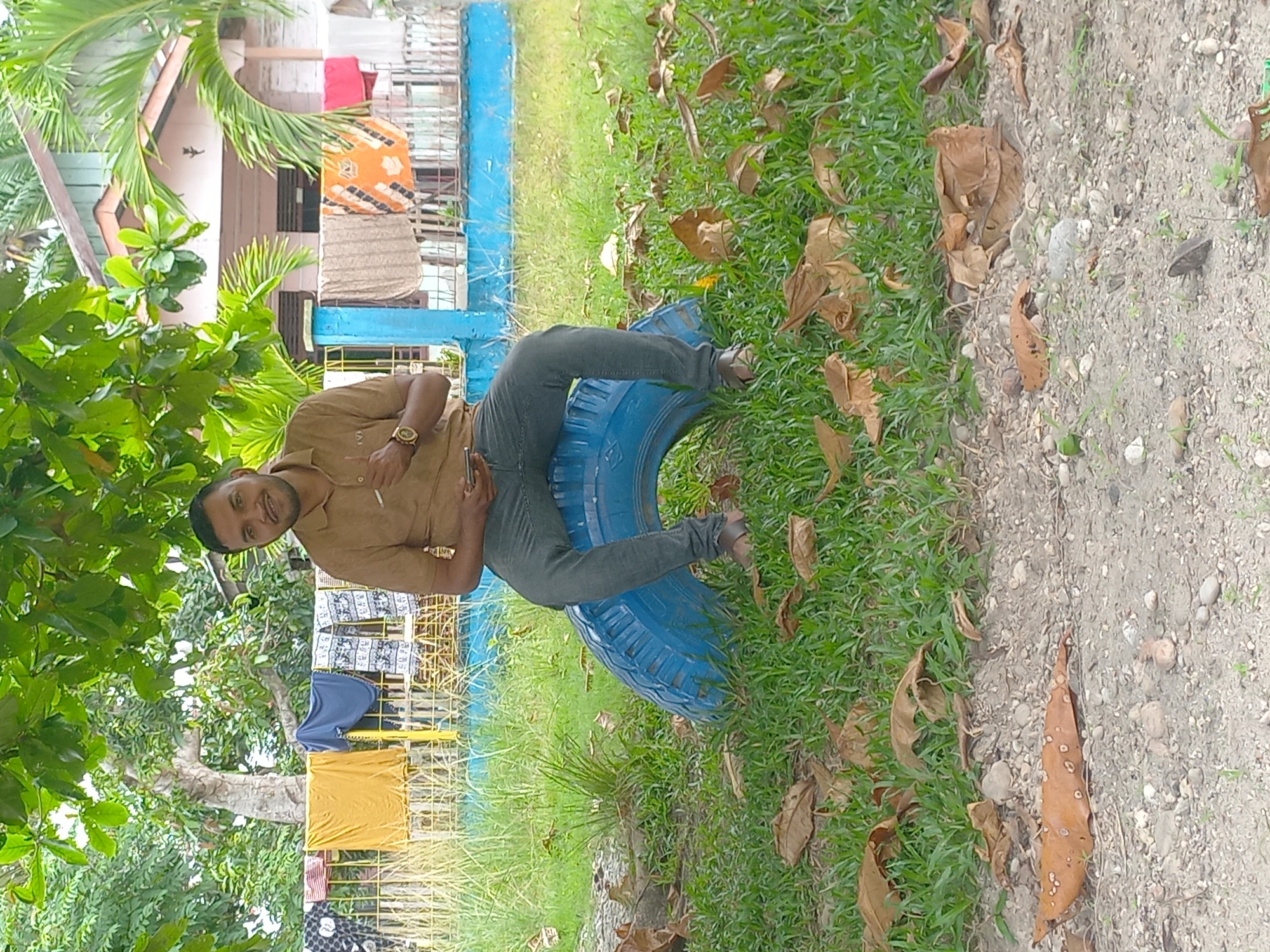 A man with dark hair and a beard is sitting on a blue tire in an outdoor setting in Gampongkeude, Indonesia. He is wearing a tan short-sleeved shirt and dark pants. His left hand is resting on his lap, holding a phone, and his right hand is extended, holding a small object, possibly a cigarette. Behind him, a fence with clothes hanging on it is visible, along with a house with a light-colored exterior and a darker roof. Palm trees and other greenery are present in the background. The foreground consists of grass and fallen leaves, with a gravelly path visible at the bottom. The lighting suggests it is daytime.