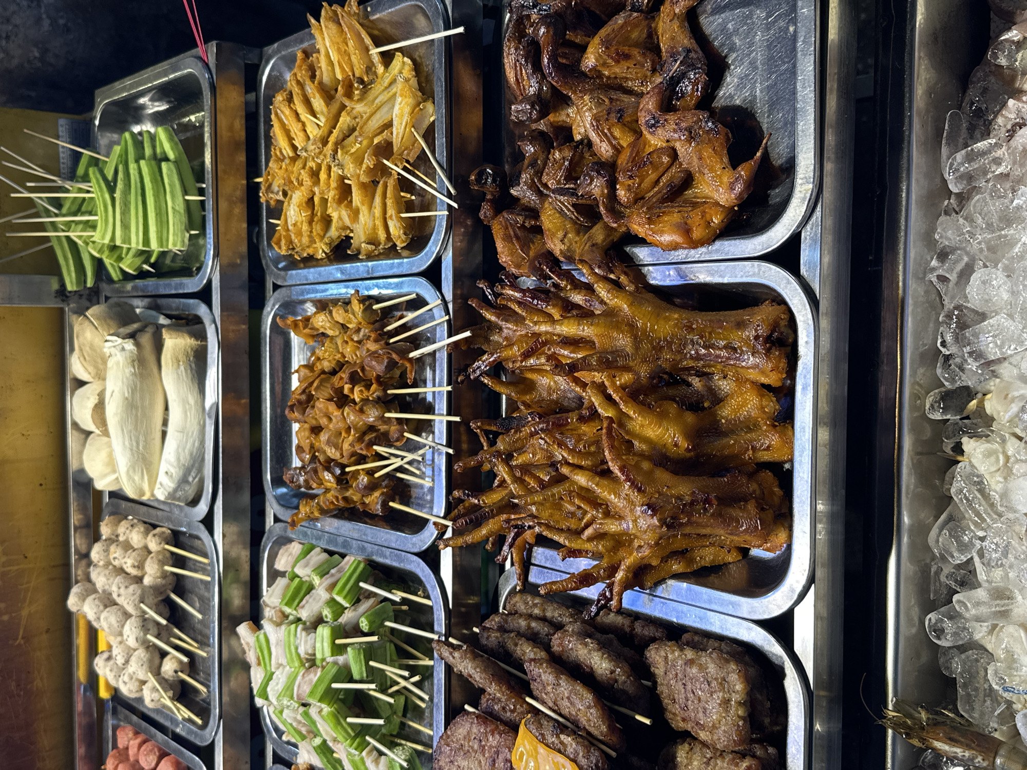This image captures a vibrant and diverse display of street food at what appears to be a night market stall in Thủ Đức, Vietnam. Various skewered food items are neatly arranged in multiple stainless steel trays, illuminated by artificial light, suggesting it is nighttime or evening.
On the left side of the frame, stacks of metal trays present a selection of vegetables and processed items. The top tray holds numerous skewers of bright green okra. Below it, a tray showcases whole, light-colored mushrooms, possibly king oyster mushrooms. Further down, a tray is filled with small, spherical meatballs on skewers, and the lowest visible tray contains skewers of reddish sausages.
The right side of the display features a variety of cooked meats. A prominent tray in the middle is piled high with golden-brown chicken feet, appearing grilled or roasted. Above this, another tray contains numerous small, whole birds, likely quail, also cooked to a deep brown. Below the chicken feet, a tray holds skewers of darker, cooked meat or offal, possibly pork or beef, with some charred edges. Adjacent to these, another tray offers skewers featuring alternating pieces of green vegetable (similar to okra) and white meat or fat.
To the far right, a tall metal container is filled with crushed ice, with some fresh seafood, specifically a prawn or shrimp, visible amongst the ice, indicating fresh ingredients are kept chilled. All the skewered items are prepared on thin wooden sticks, typical for street food. There is no visible text in the image. The overall scene reflects a common and inviting aspect of Southeast Asian street food culture.