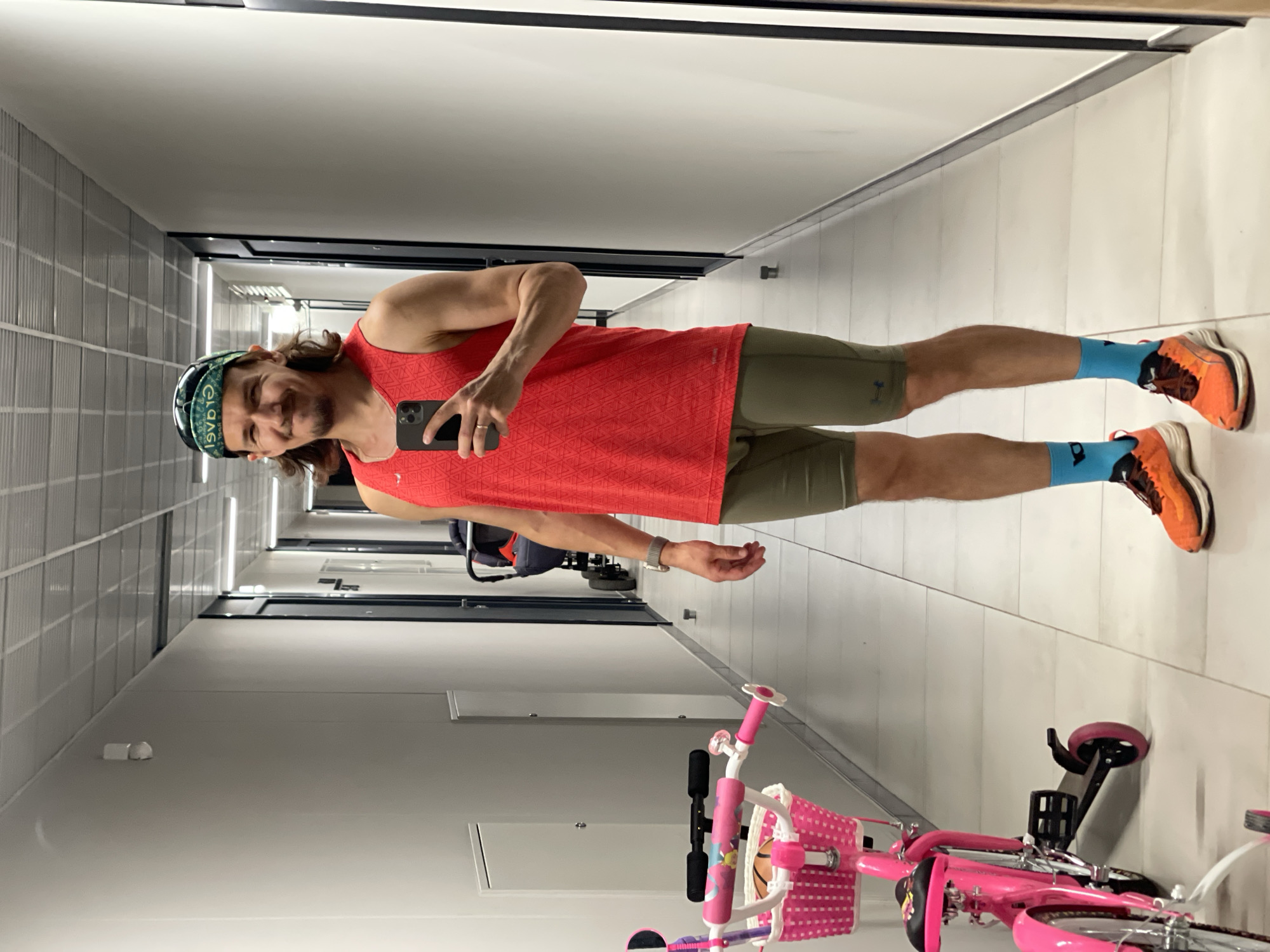 A man is standing in a well-lit, modern hallway, taking a selfie in a mirror. He is wearing a red sleeveless shirt, olive green athletic shorts, bright blue socks, and orange running shoes. He has a green and black bandana on his head and a watch on his left wrist. His right hand holds a black smartphone, and a ring is visible on his left ring finger. To his left, a pink child's bicycle with a basket containing a basketball is parked. In the background, a stroller is visible further down the hallway. The walls are white, and the floor is tiled in a grid pattern. The ceiling features recessed lighting and a metallic grid.