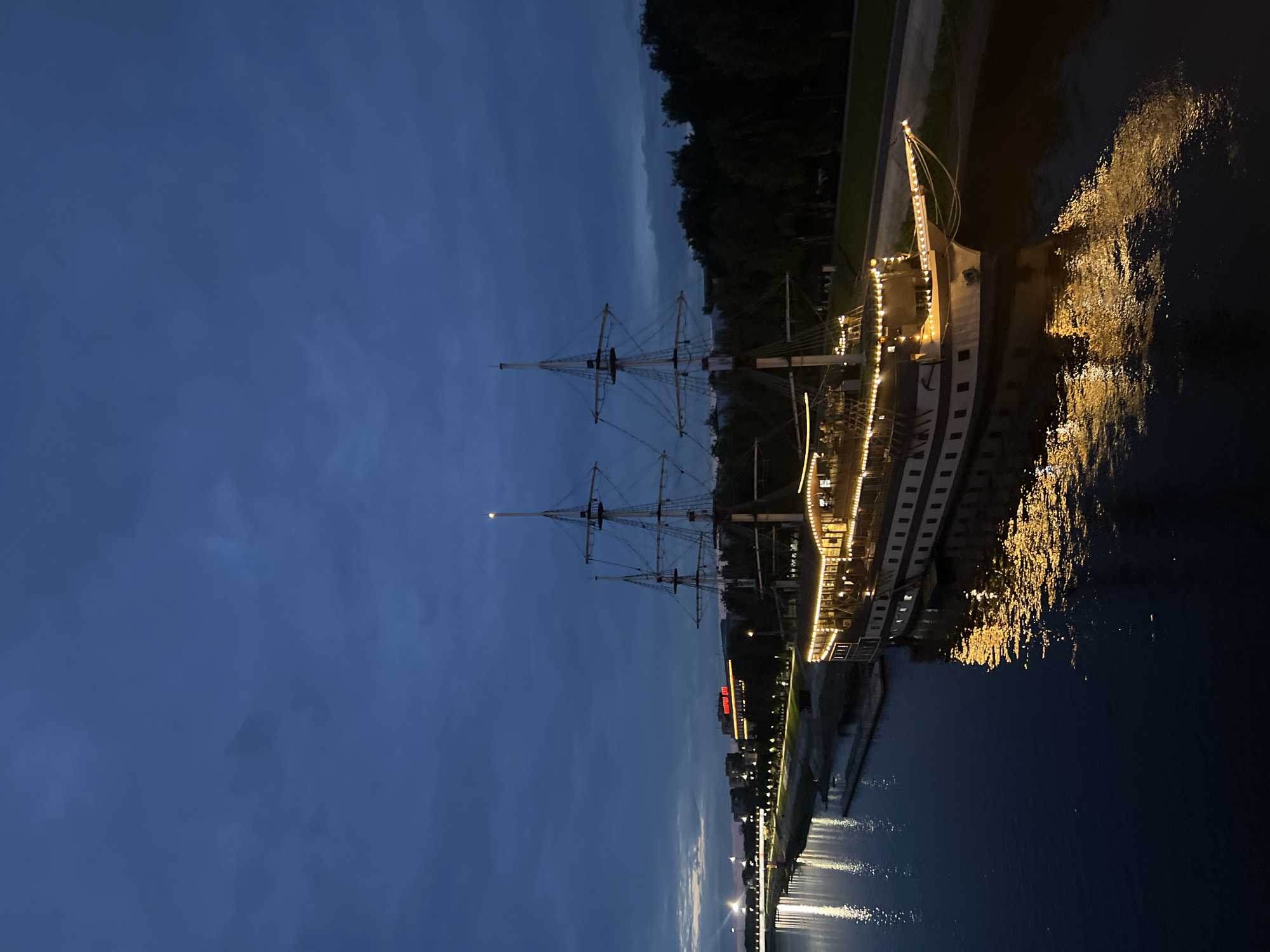 A large, illuminated sailing ship with three masts is docked on a riverbank at dusk in Veliky Novgorod, Russia. The ship's hull is white and dark, and it is adorned with numerous yellow lights along its deck and railings. The water reflects the ship's lights and the lights of a bridge in the distance. Across the river, a city skyline is visible with buildings and trees, including a building with a red illuminated sign. The sky is dark blue with some clouds.
