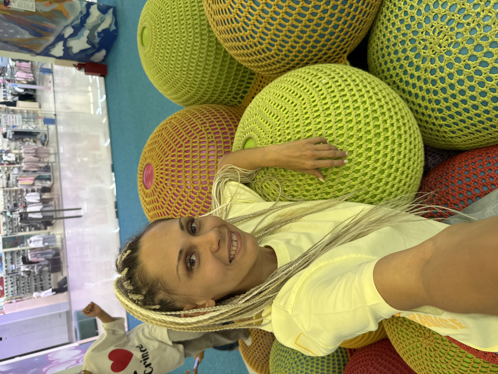 A woman with blonde braided hair is taking a selfie in an indoor public space, possibly a mall or play area, with a turquoise floor. She is wearing a yellow t-shirt. She is sitting or leaning against several large, colorful, crocheted poufs. In the background, there are shops with clothing displays visible through glass. To the right, there is a mural of a mountain landscape. The exact location in the city of Zhukovo, Russia is not specified in the image.