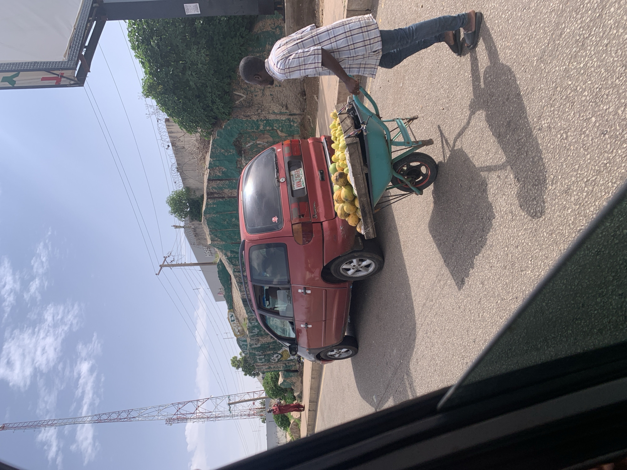 A person is pushing a wheelbarrow filled with fruits, possibly mangoes, alongside a red minivan. The scene appears to be a street in Jos, Nigeria. The person, wearing a plaid shirt and jeans, is pushing the wheelbarrow from behind, with their right hand on the handle. The wheelbarrow is a bright teal color and has a wooden platform to hold the fruit. The minivan is parked on the right side of the road, and its rear is facing the viewer. The wheelbarrow is positioned to the right of the minivan.
The ground is asphalt, suggesting a paved road. There are trees and some fencing visible in the background, along with power lines and a tall, red and white structure that resembles a communication tower or a bridge. The sky is blue with some scattered white clouds, indicating it is daytime. The shadows cast by the person and the wheelbarrow suggest the sun is at an angle, likely mid-morning or afternoon.
A license plate is visible on the back of the minivan, with the text "AN K2644". A sign is partially visible on the left side of the image with text that is too blurry to read. The overall impression is a common street scene in a bustling urban environment, with a vendor likely transporting their goods.