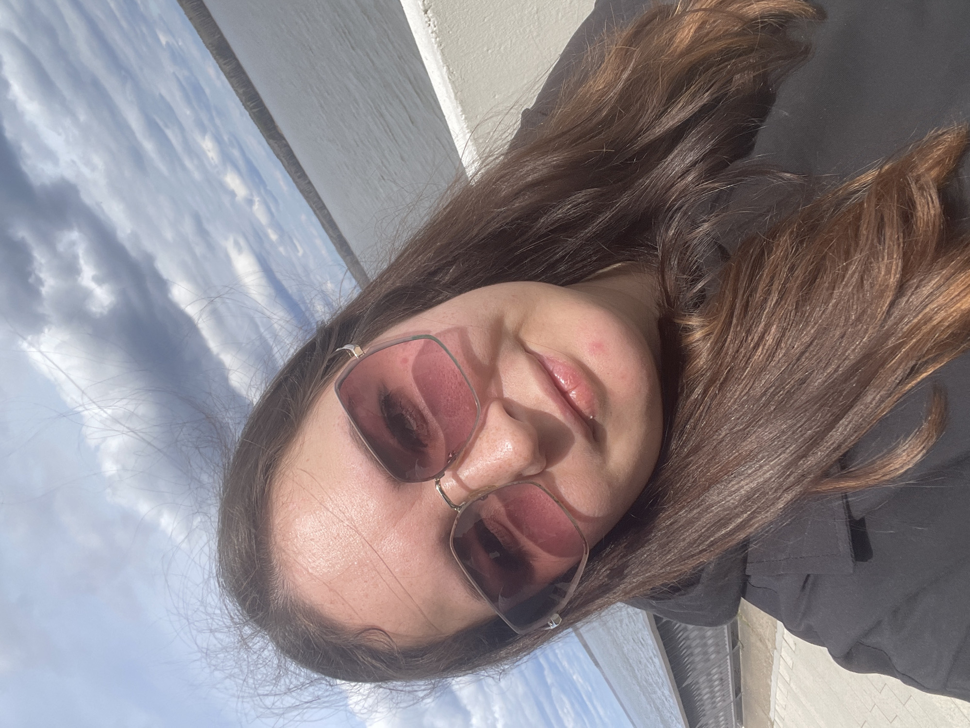 A young woman with long, brown hair and sunglasses is the focus of the image. She is wearing a black jacket. The background shows a sky with light clouds and a body of water. The photo appears to have been taken outdoors during the daytime. While the location cannot be definitively determined, the presence of what appears to be a large river or reservoir suggests it is likely a location near water with open skies. Without explicit landmarks, it's impossible to confirm if this image was taken in Chaikovsky, Russia.