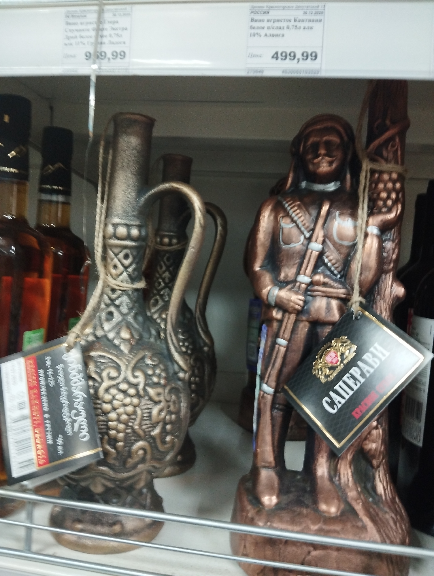 Here is a forensic description of the image:
**Scene Overview:** The image depicts a shelf in a store, likely in Krasnogorskoye, Russia, based on signage mentioning "Россия" (Russia). The shelf contains decorative bottles and a statuette of a male figure.
**Objects:**
* **Bottles:** There are several bottles of alcohol, possibly wine and brandy or whiskey, visible on the shelf.
* **Decorative jugs:** There are two clay or ceramic jugs with decorative designs, possibly representing grapes, and handles. They have tags attached to them that read “ქინძმარაული”.
* **Statuette:** A brown, bronze-colored statuette of a male figure holding a rifle and grapes, possibly a soldier or hunter. It has a tag attached that reads “САПЕРАВИ”.
* **Price Tags:** Price tags are visible on the shelf, with amounts listed as "969,99" and "499,99".
**Environment:**
* **Setting:** The setting is a retail environment, specifically a store aisle.
* **Lighting:** The lighting appears to be artificial, typical of a store setting.
* **Shelving:** Items are placed on metal wire shelving.
**Details from Text on Tags/Signs:**
* A sign states the product is "Вино игристое Кантиани" (Kantian Sparkling Wine), "белое п/слад 0,75л" (white semi-sweet 0.75L), with "10% Алвиса" (10% Alvisa).
* "ქინძმარაული"
* "САПЕРАВИ"