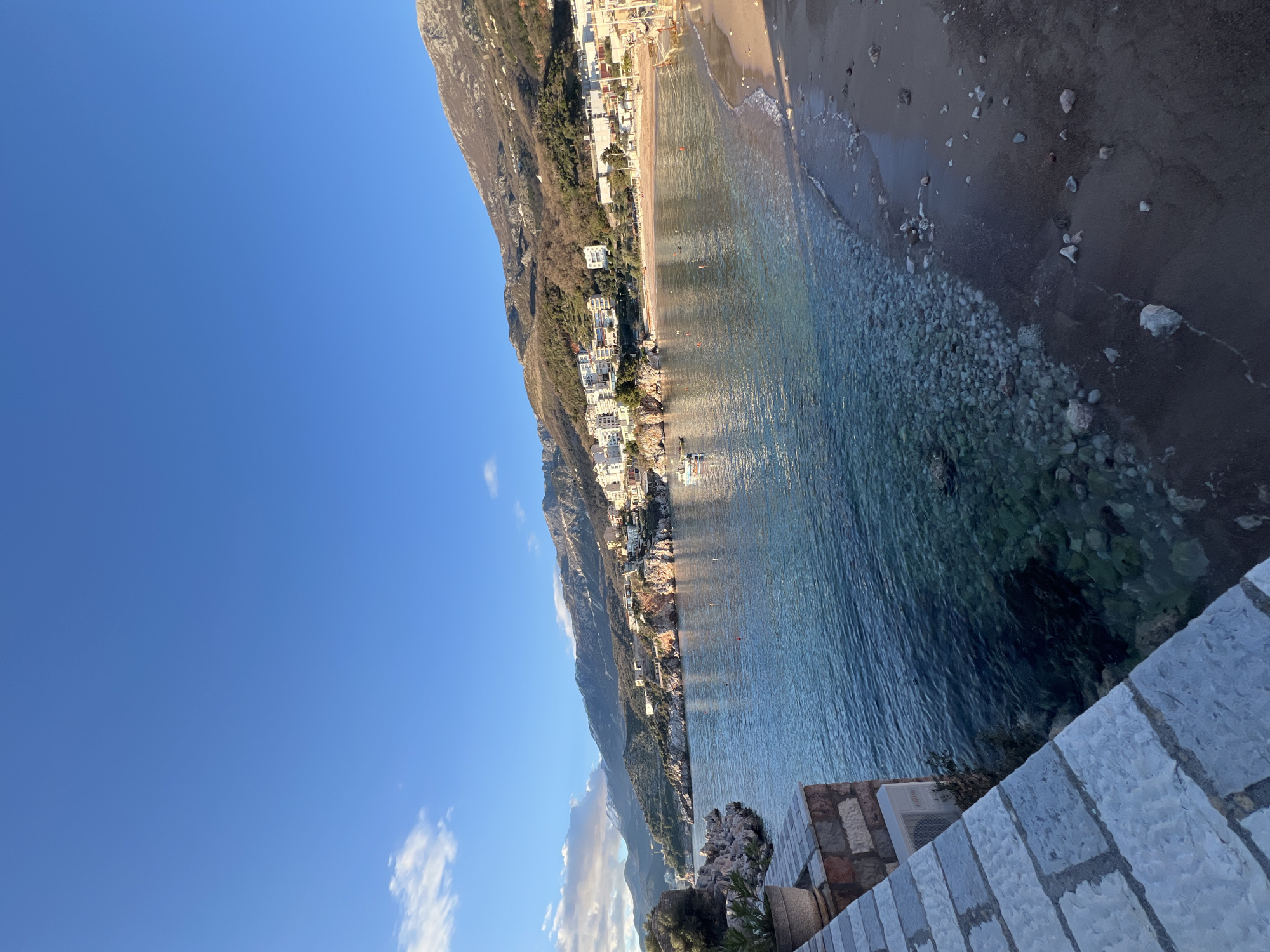 The image captures a coastal view in Boreti, Montenegro. The Adriatic Sea occupies the center, transitioning from turquoise near the shore to deeper blue further out. The shoreline is a mix of sand and pebbles, with scattered rocks along the water's edge. A mountain range rises sharply alongside the coast, with buildings clustered along its lower slopes and the waterfront. The sky is a clear, bright blue, with light clouds visible towards the bottom left of the frame. A stone wall and some vegetation appear in the foreground. No individuals are clearly identifiable.