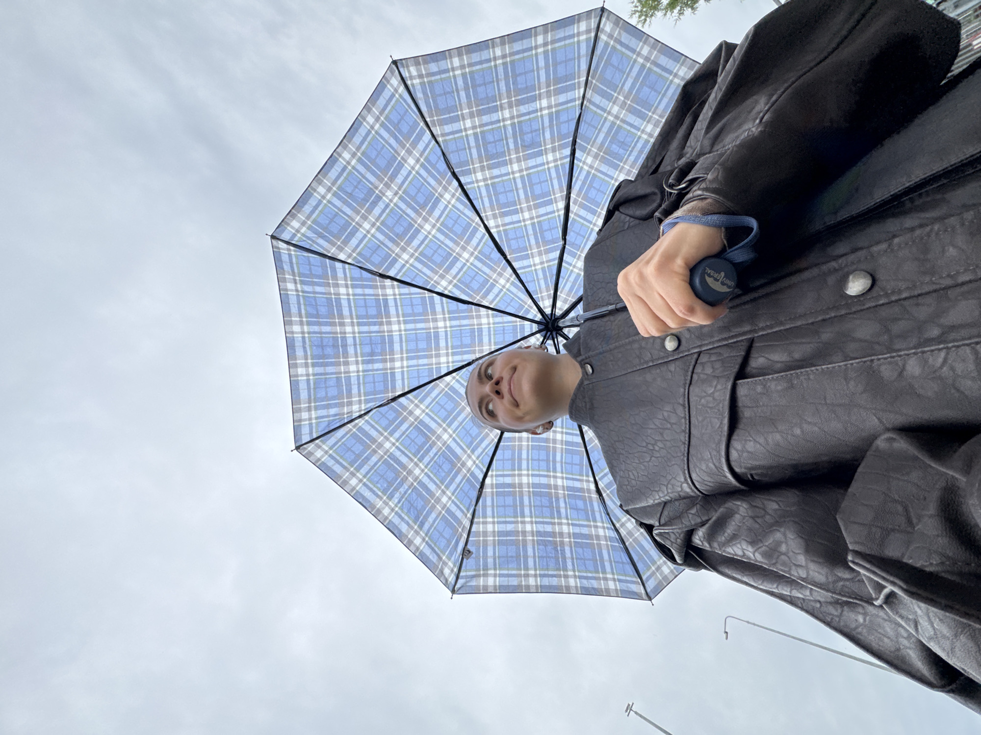 The image is an extreme low-angle shot of a person holding a blue and white plaid umbrella open above them. The person is wearing a dark-colored, textured jacket, possibly leather. Their head is tilted back, looking up towards the umbrella and the sky. The umbrella dominates the upper portion of the frame, with its spokes and fabric clearly visible. The sky is overcast and appears cloudy. The lighting suggests it is daytime, possibly a slightly gloomy or rainy day, given the umbrella. The overall setting appears to be outdoors, with a hint of what might be streetlights or poles in the distance at the bottom of the frame. The person's expression is difficult to discern fully due to the angle, but they seem to be looking upwards with a neutral or slightly curious expression. The focus is on the person and the umbrella, with the sky as the backdrop. There is some visible text on the handle of the umbrella, which reads "UNIVERSAL".