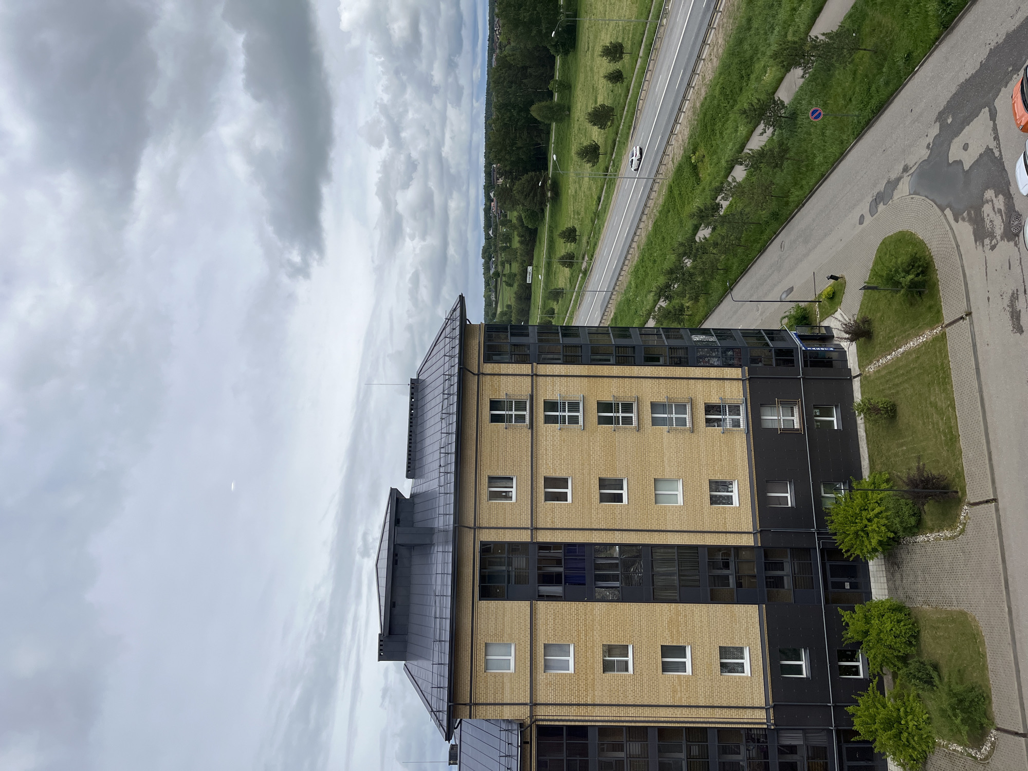 View of an urban landscape featuring a modern building with a brick facade, set against a backdrop of cloudy skies and a tree-lined highway. The building has a mixed-use architectural style, with a combination of yellow brick sections and dark glass or metal panels. The surroundings include a paved area with small landscaped patches and a view overlooking a roadway with sparse traffic and a green belt with scattered trees. Based on the implied location from the image request, the scene is indicative of Мокшино, Russia.