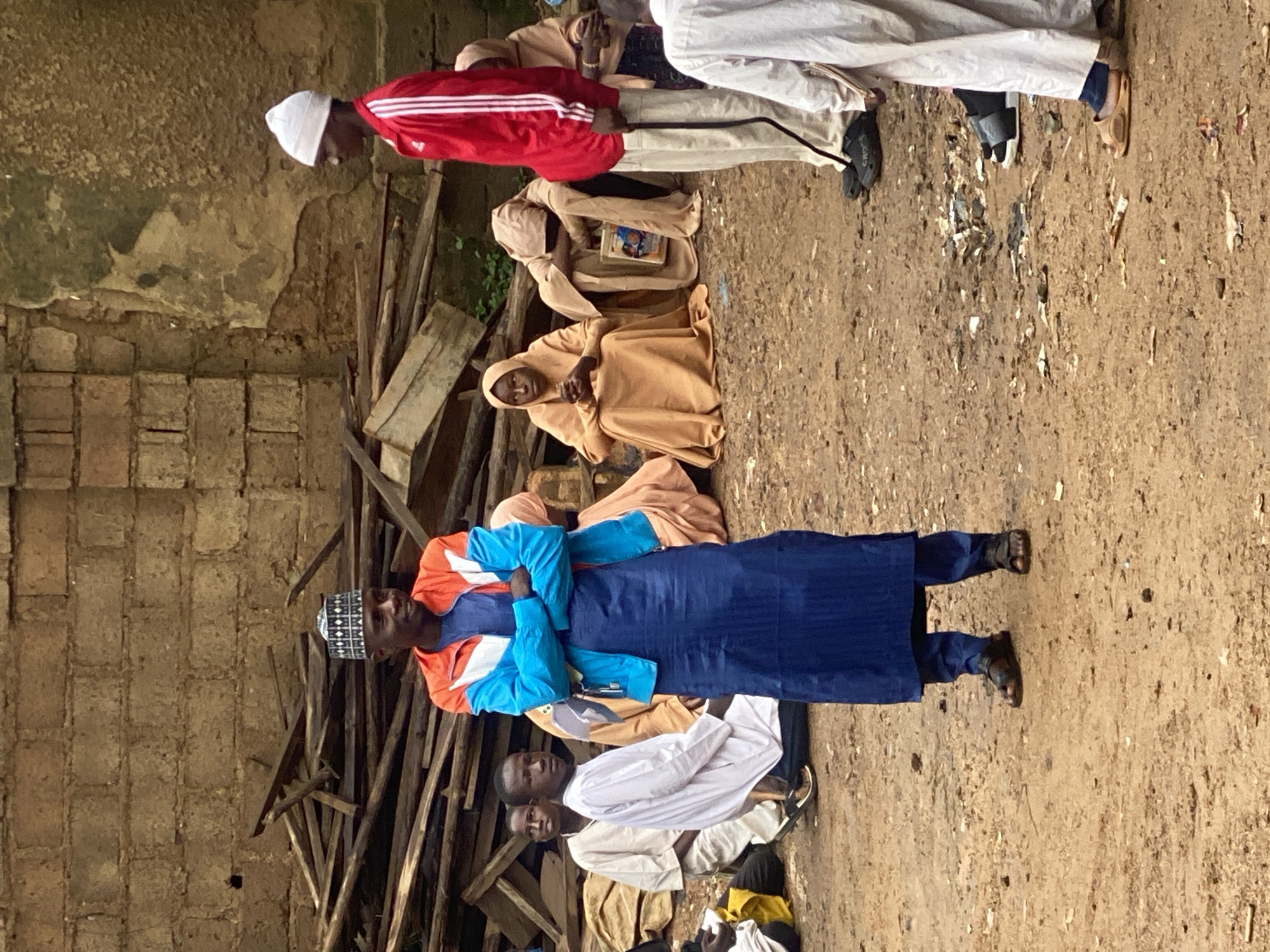 In an outdoor environment in Jos, Nigeria, a group of people are gathered on dirt ground. A man in the foreground is wearing a blue robe and an orange and blue jacket, with his arms crossed. To his right, another man in a red jacket and white beanie stands facing away from the camera. Behind them, several people are seated, some wearing light brown or tan robes and headscarves. A pile of wooden planks is visible in the background against a brick wall. The scene appears to be in an open, somewhat unkempt area, possibly a courtyard or a less developed part of the city.