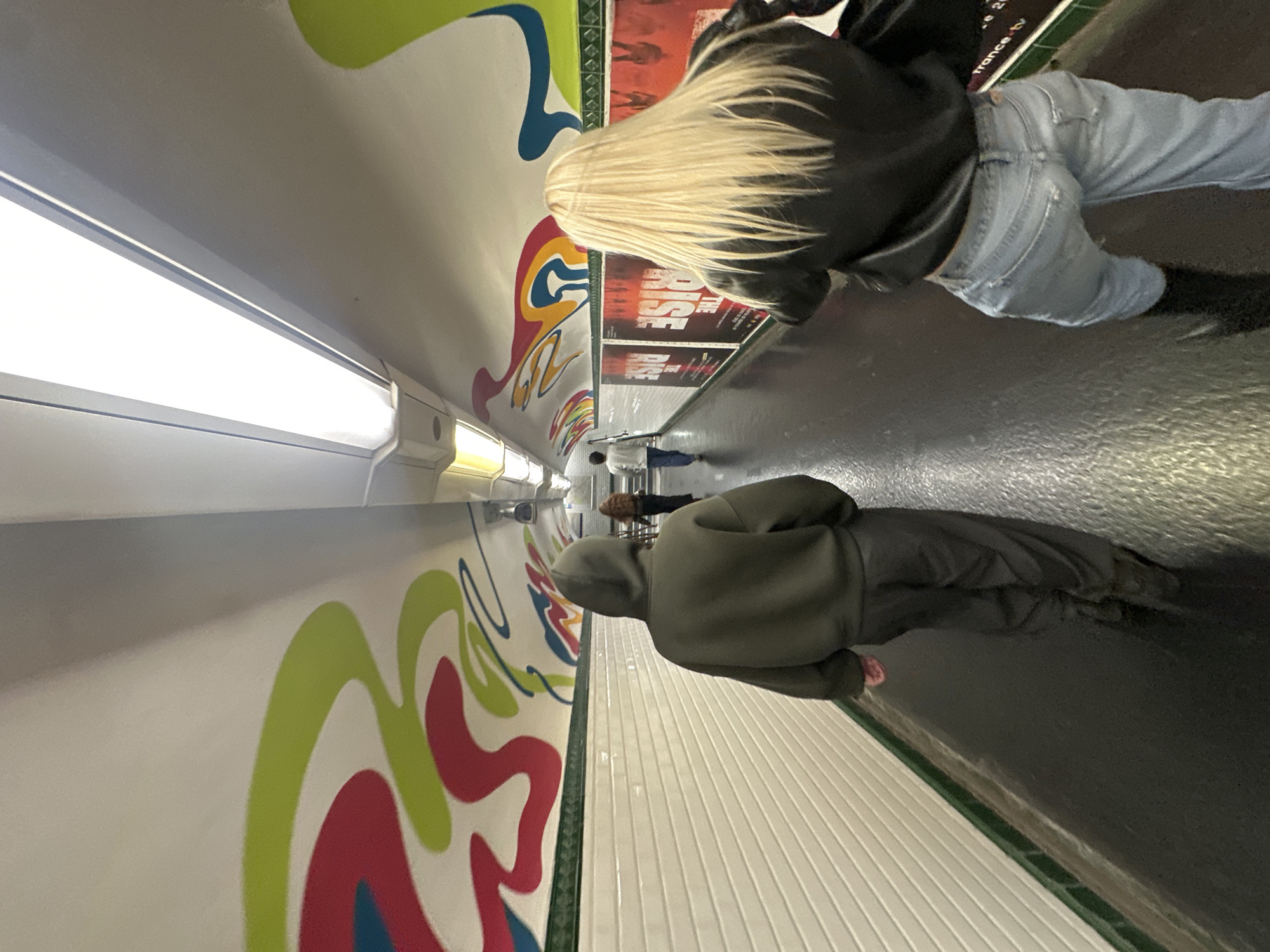 The image shows the interior of a Paris Metro car. A white wall with psychedelic, swirling patterns of red, green, and blue extends along the left side. Above this wall are fluorescent lights. Above this is a gray ceiling. A row of advertising posters runs along the upper right side of the image.
Several people are visible in the center of the frame. Two people are standing closer to the camera. One person wears a green hoodie. Another person stands further from the camera wearing a dark jacket and light-wash denim. Several figures are seen further down the metro car.
The floor is dark and gray. The walls are white with green trim and lined with horizontal ridges.