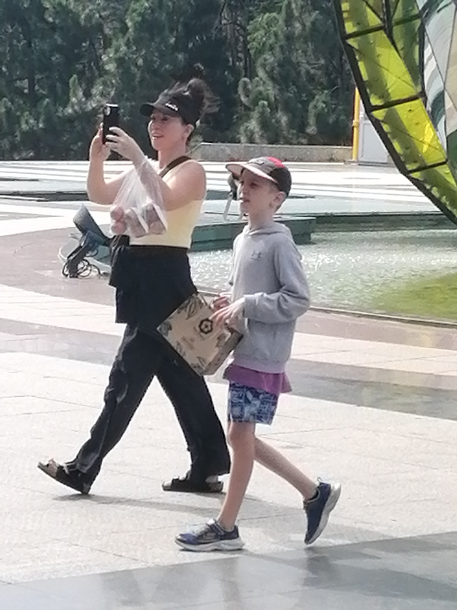 An adult woman and a young boy are walking side-by-side on a paved surface outdoors. The woman, on the left, is wearing a yellow tank top, black pants, and dark sandals. She has a black baseball cap on backwards and is holding a clear plastic bag containing items and a smartphone in her left hand, appearing to take a photo. A bag is slung over her right shoulder. The boy, on the right, is wearing a grey hooded sweatshirt, a purple undershirt, and patterned shorts. He has a baseball cap with red accents on his head and is wearing blue athletic shoes. He is holding a brown paper bag in his hands. The background features trees and a body of water. The scene does not appear to be in Da Lat, Vietnam; it is a generic outdoor setting.