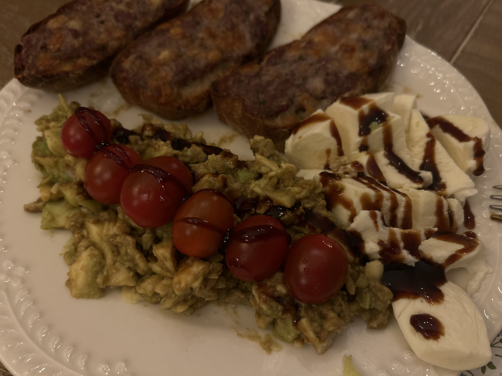 A white plate contains three toasted baguette slices topped with a dark topping, a bed of mashed avocado with six cherry tomatoes drizzled with balsamic glaze, and sliced fresh mozzarella drizzled with balsamic glaze. The plate rests on a wooden surface. The scene likely occurs indoors, but its precise location in Plovdiv, Bulgaria cannot be determined from the image.