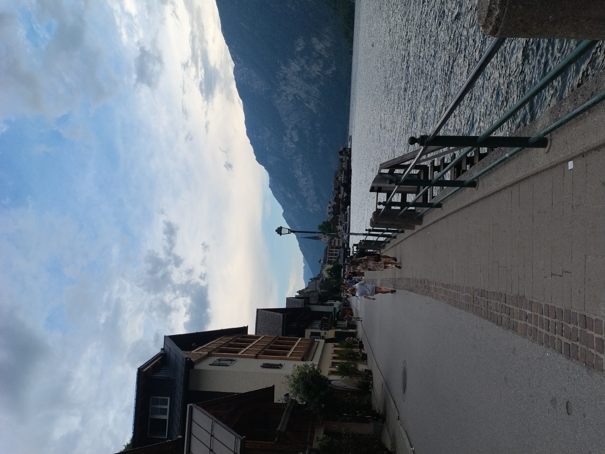 The image depicts a view of the waterfront in Hallstatt, Austria, under a cloudy sky. On the left, a row of traditional European buildings lines a paved street. The architecture features dark roofs and multi-story facades with wooden accents. Several people are walking along a pedestrian walkway adjacent to the street, moving away from the camera. To the right of the walkway, a dark metal railing with wooden elements separates the path from a wide body of water. In the background, across the water, a mountain range rises steeply, with a small cluster of buildings nestled at its base, including a prominent church spire. The water is a deep blue-grey, reflecting the overcast sky and the surrounding landscape. The overall scene conveys a serene, albeit slightly somber, atmosphere typical of a European lakeside town.