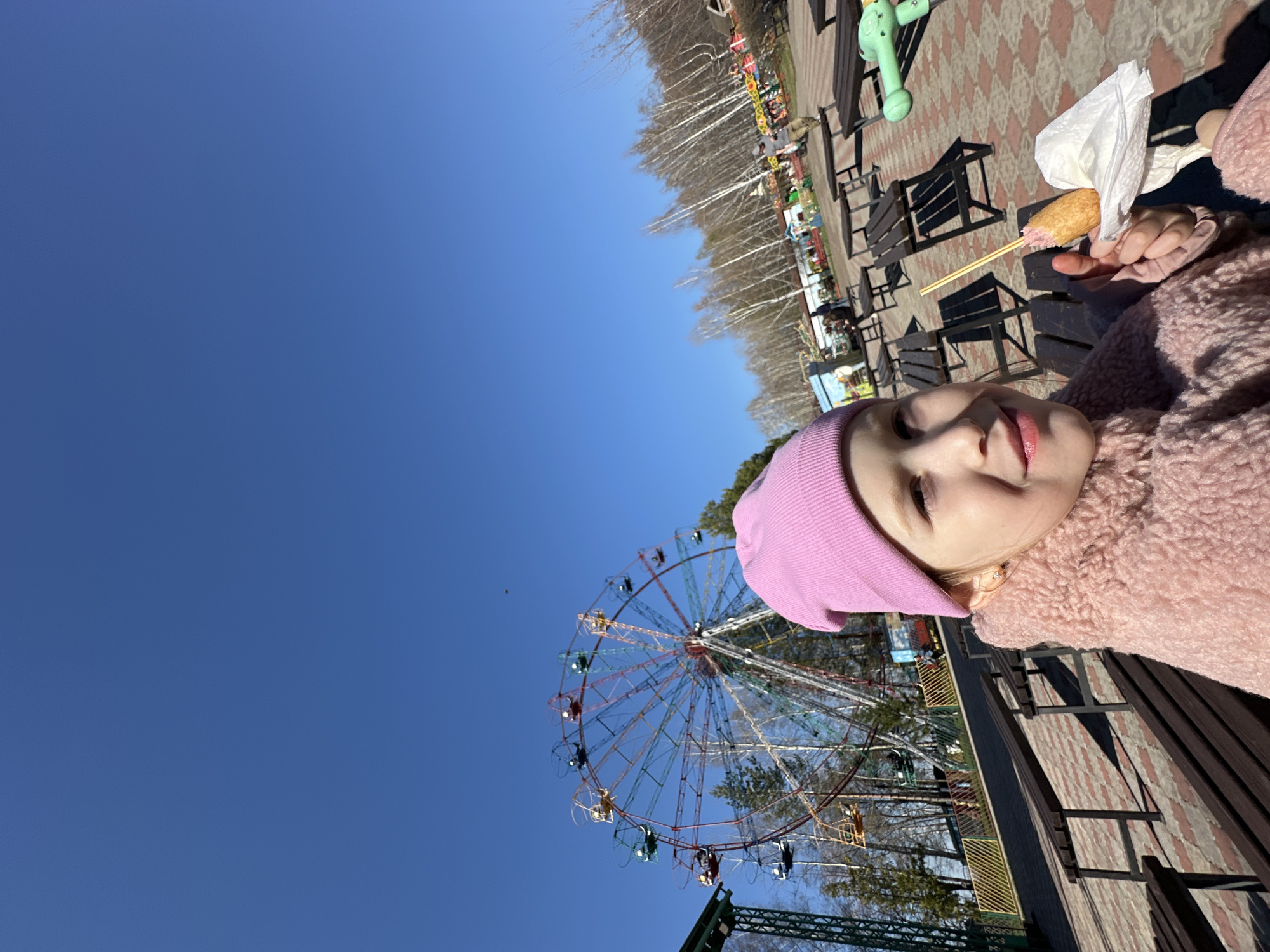 A young girl in a pink hat and pink fuzzy jacket is holding a corn dog with a white napkin in her right hand. She is smiling slightly. Behind her is a Ferris wheel with brightly colored cars in Chaikovsky, Russia. The sky is bright blue and clear. There are benches and trees in the background. The ground is paved with a reddish-brown and white patterned brick.