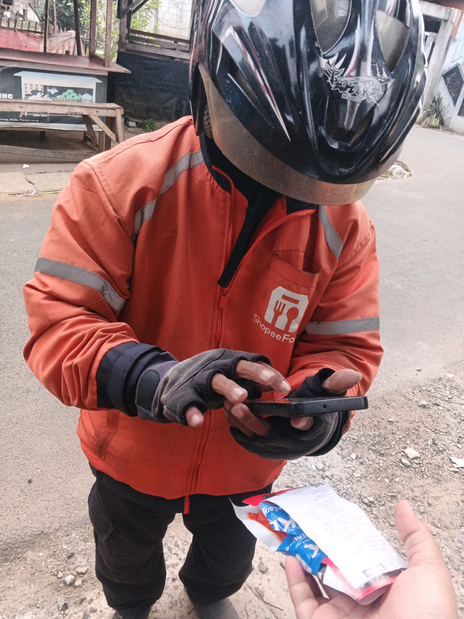 A person wearing an orange jacket with reflective stripes and black gloves is holding a smartphone and interacting with it. The jacket has a ShopeeFood logo on it. The person is wearing a black helmet. The scene appears to be outdoors, on a road with gravel and asphalt. In the foreground, another person's hand is offering a receipt or slip of paper, which contains some printed text and what appears to be packaging for a snack or food item. The background shows some wooden structures and vegetation, indicative of an urban or semi-urban environment. The location is identified as Kadu, Indonesia.