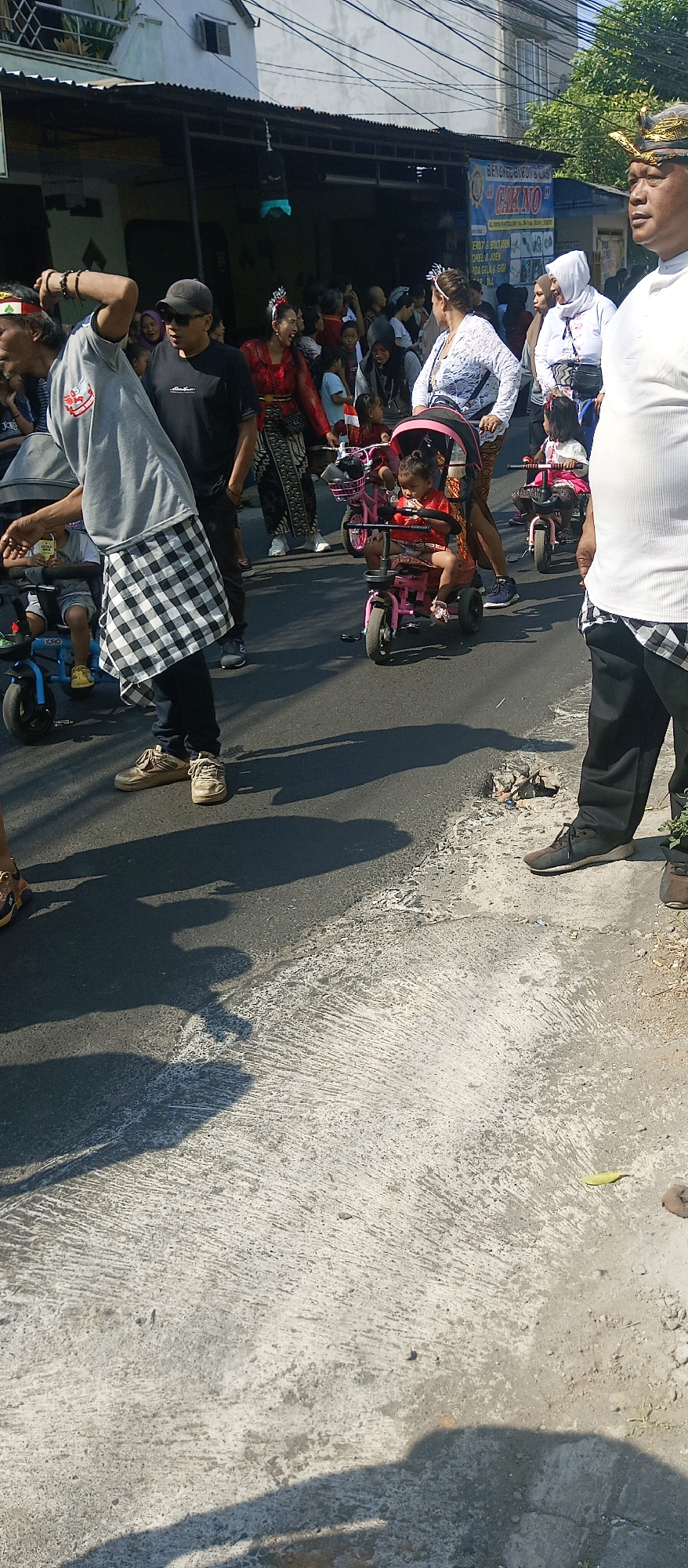 This image depicts a street scene in Malang, Indonesia, likely during a parade or festival. Several people are visible, including adults and children. In the foreground, a man in a grey t-shirt and checkered cloth tied around his waist stands with his back to the camera. To his right, a child sits in a pink tricycle, looking down. Further to the right, another child is in a similar pink tricycle, and an adult woman in white attire is pushing it.
In the background, more people are gathered, some wearing traditional clothing, including a woman in a red outfit with elaborate headwear. A building with a dark awning runs along the background. A banner with Indonesian text, including "CAK NO," is visible hanging from the awning. The ground is asphalt with some concrete and debris. The lighting suggests it is daytime, with strong shadows cast by the individuals. The presence of tricycles and the celebratory attire hints at a community event or procession.