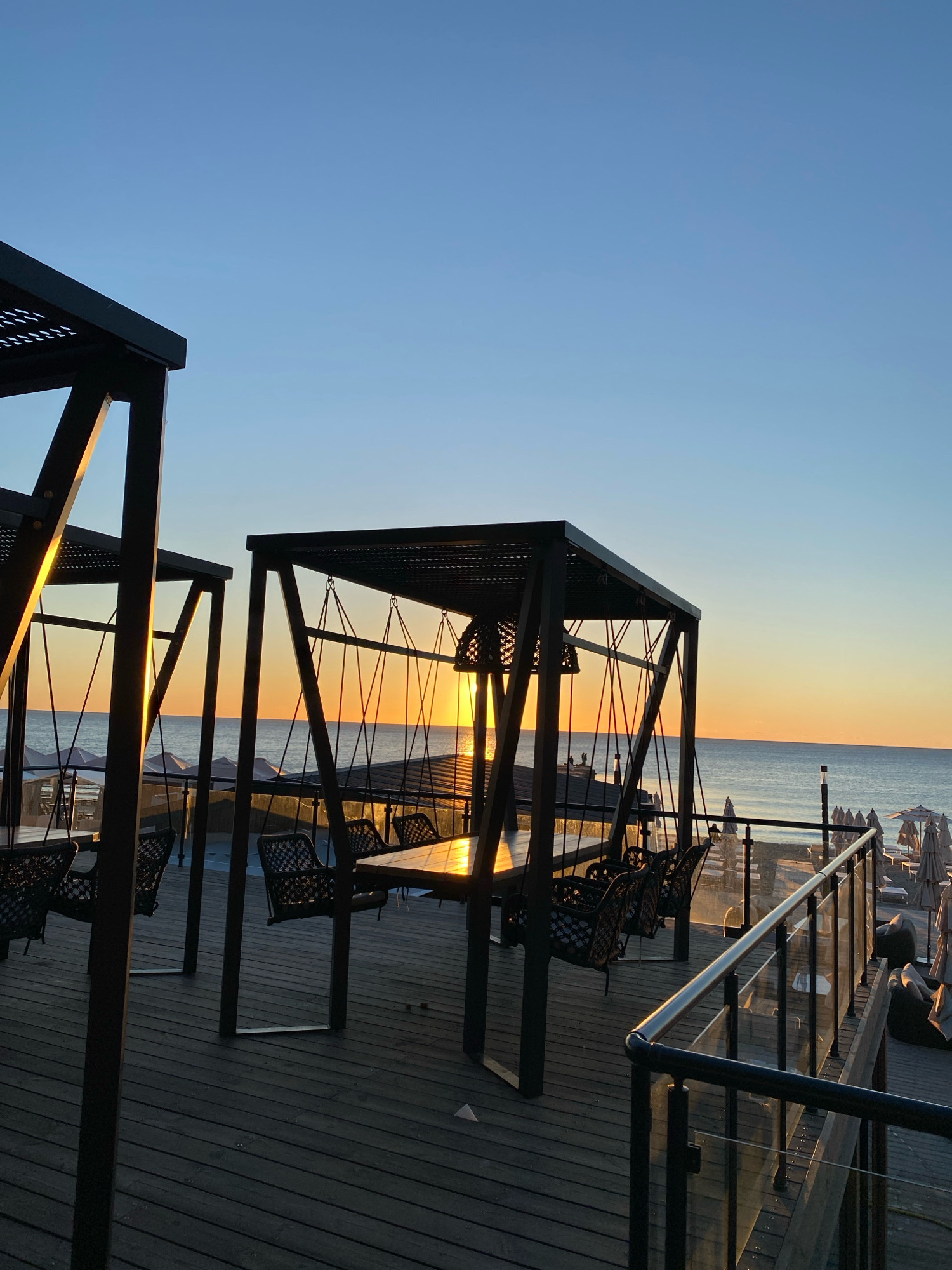 The image depicts an outdoor seating area with swings and tables on a wooden deck, overlooking the Black Sea at sunrise. The scene is set in Odesa, Ukraine. The sky is a gradient of deep blue to pale orange, indicating early morning. The sun is just above the horizon, casting a warm glow on the water and the structures. Two black metal pergolas, each housing swings and tables, are prominent in the foreground. The swings are made of dark woven material. In the background, beach umbrellas and lounge chairs are visible on a sandy shore. A glass and metal railing separates the deck from the lower level of the beach. There are no people visible in the image.