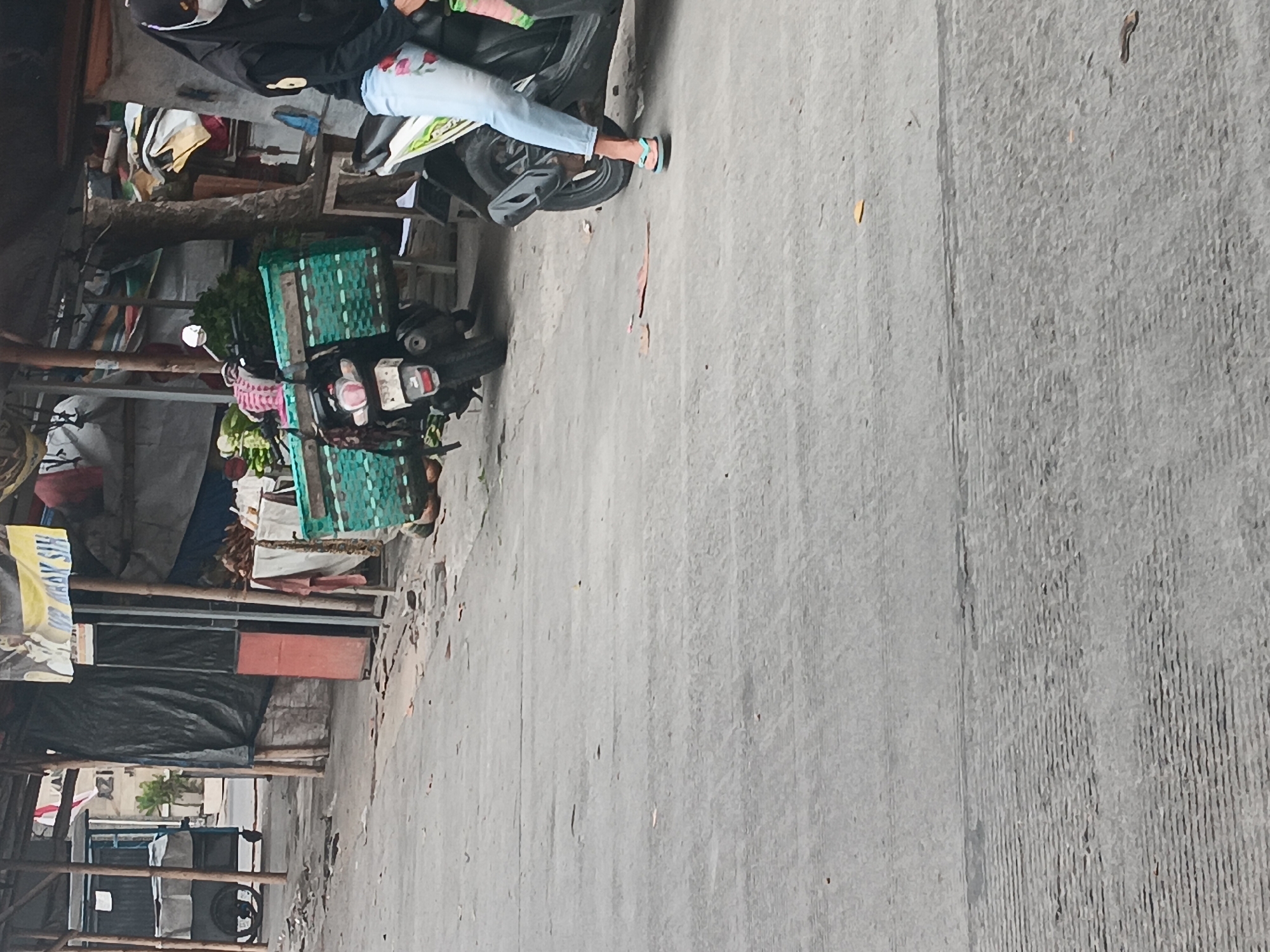 A medium shot, taken outdoors in Batursari, Indonesia, shows a roadside market scene. In the foreground, a grey asphalt road is visible, with some scattered leaves and debris. To the right, a woman wearing a black headscarf, mask, black jacket, and light blue jeans with a floral pattern is seated on a white scooter. She is wearing flip-flops. The scooter has a green, woven basket attached to the back, containing produce, possibly vegetables. To the left of this scooter, another scooter is partially visible, also fitted with a similar green basket carrying goods. Behind the scooters, market stalls are set up with tarps and wooden structures. One stall has a cart with white coverings. The background reveals more market activity and structures, including a red cylindrical object. The overall impression is that of a bustling, everyday scene at a local market.