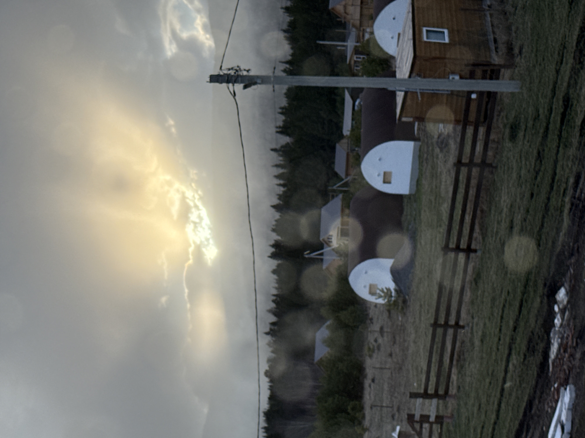 The image shows an outdoor scene, likely in a rural area, possibly near the city of Tyulyuk, Russia. A wooden building with a window is visible, adjacent to a wooden fence. There are also white, half-dome structures with square openings. A power line runs vertically through the image, supported by a utility pole. The background features a line of evergreen trees. The sky is overcast with a patch of bright sunlight. The image appears to have been taken through a window or lens with water droplets.