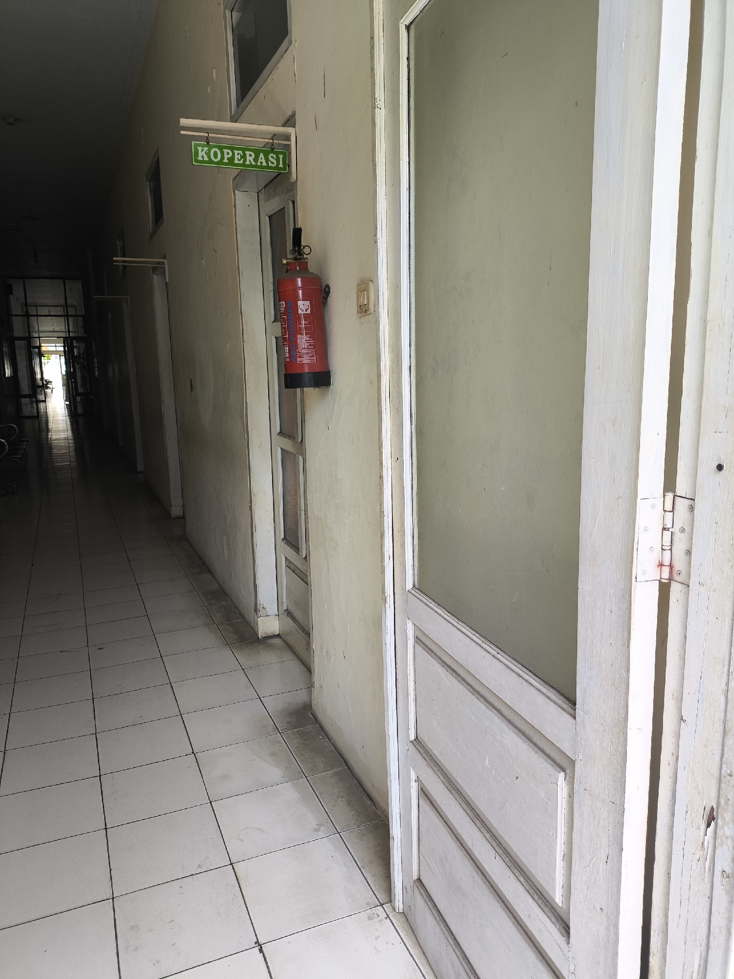 The image depicts a dimly lit hallway with tiled flooring in what appears to be an institutional building. On the left side of the hallway, a green sign with white lettering reading "KOPERASI" is affixed to the wall above a door. To the right of the sign, a red fire extinguisher is mounted on the wall. The hallway recedes into the distance, showing multiple doors along the walls and a brightly lit opening at the far end. The overall impression is one of a functional, perhaps slightly dated, interior space. The specific location in Cilegon, Indonesia, cannot be determined from the visual information alone.