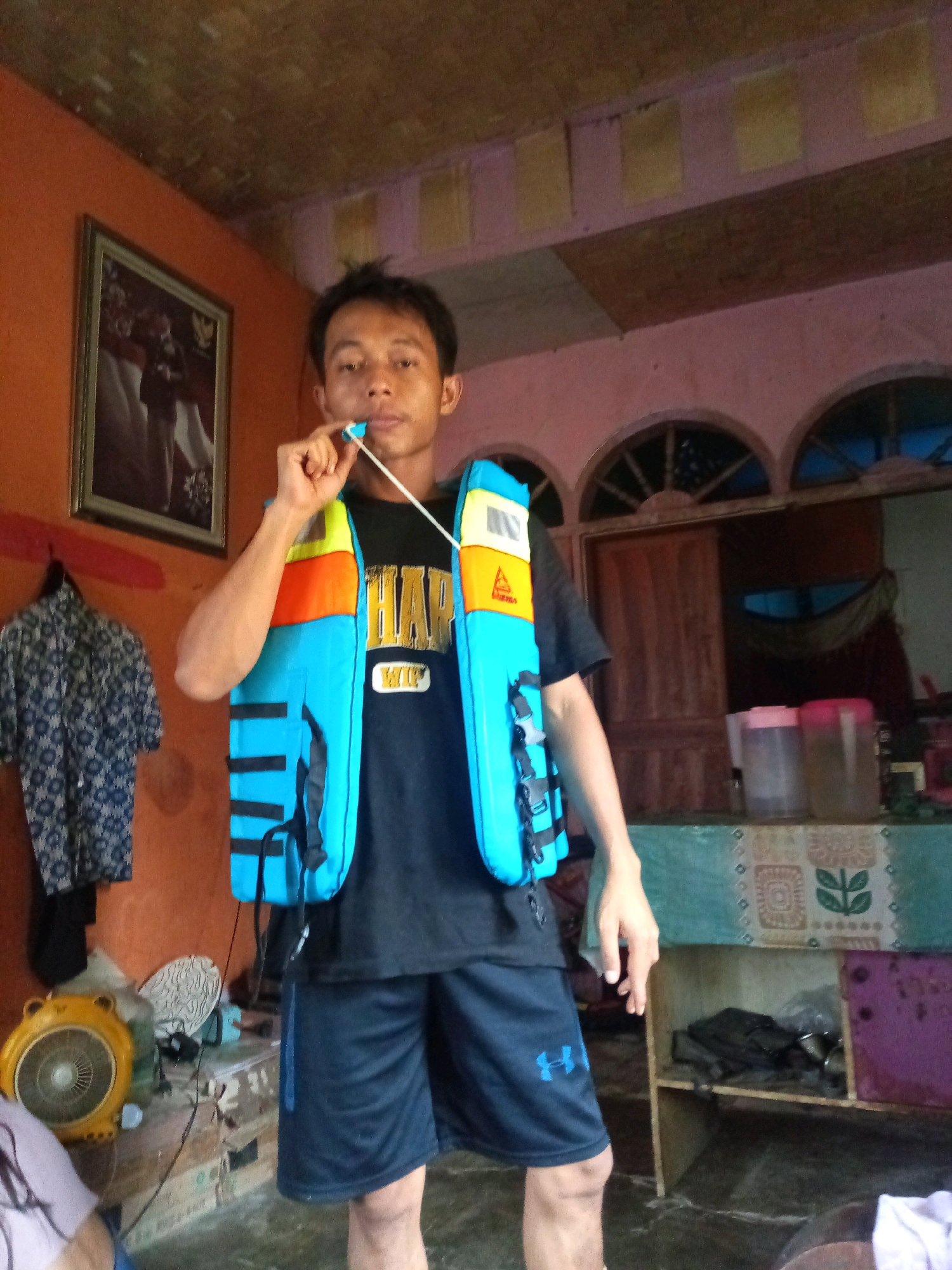 A young man stands indoors, holding a white string attached to a blue and orange life vest that he is wearing over a black t-shirt. The man has dark hair and is looking towards the camera, with the string in his mouth. The life vest is bright blue with orange and yellow accents. The t-shirt has a yellow graphic on it that is partially obscured by the life vest. He is also wearing dark blue athletic shorts. In the background, there is a wall painted orange on the left, and a pink wall on the right. A framed picture hangs on the orange wall. A patterned shirt is hanging on a hook to the left of the man. A yellow fan is on the floor to the man's left. The floor appears to be concrete or tile. There are shelves in the background to the right, holding various objects including two large plastic containers and a dark bag. The ceiling is visible at the top of the image and is made of wood. The overall impression is a candid shot taken in a casual indoor setting.