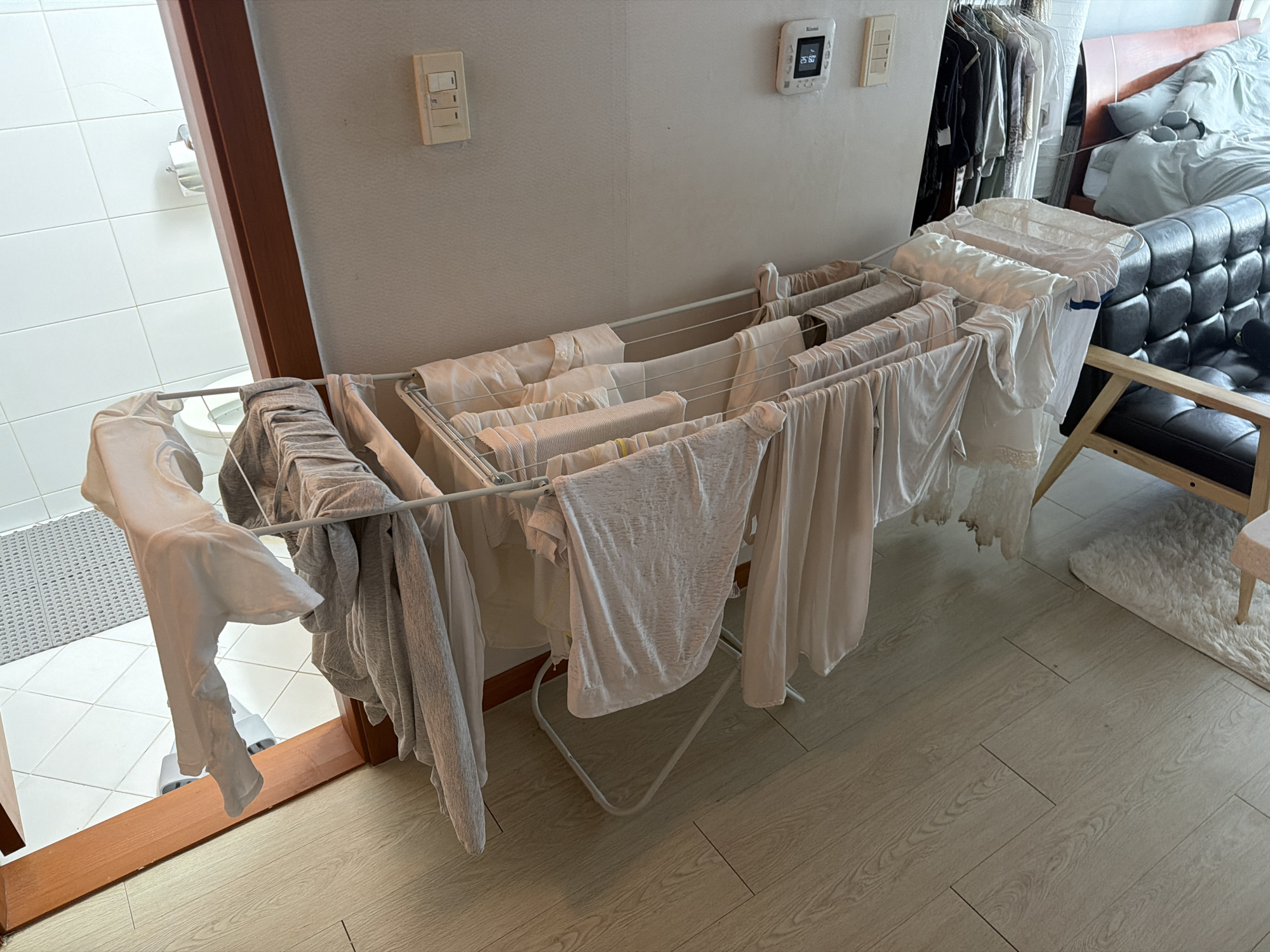 A drying rack filled with clothes dominates the view. The rack is situated in an indoor space with a tiled bathroom visible through an open doorway on the left. The interior space has wood-patterned flooring. A wall with a light switch and a digital thermostat is in the background. A closet is on the right, next to a black leather sofa and a small rug. The location is likely a residential area in Seoul, South Korea, given the style of the interior design and the type of thermostat. No people are visible.