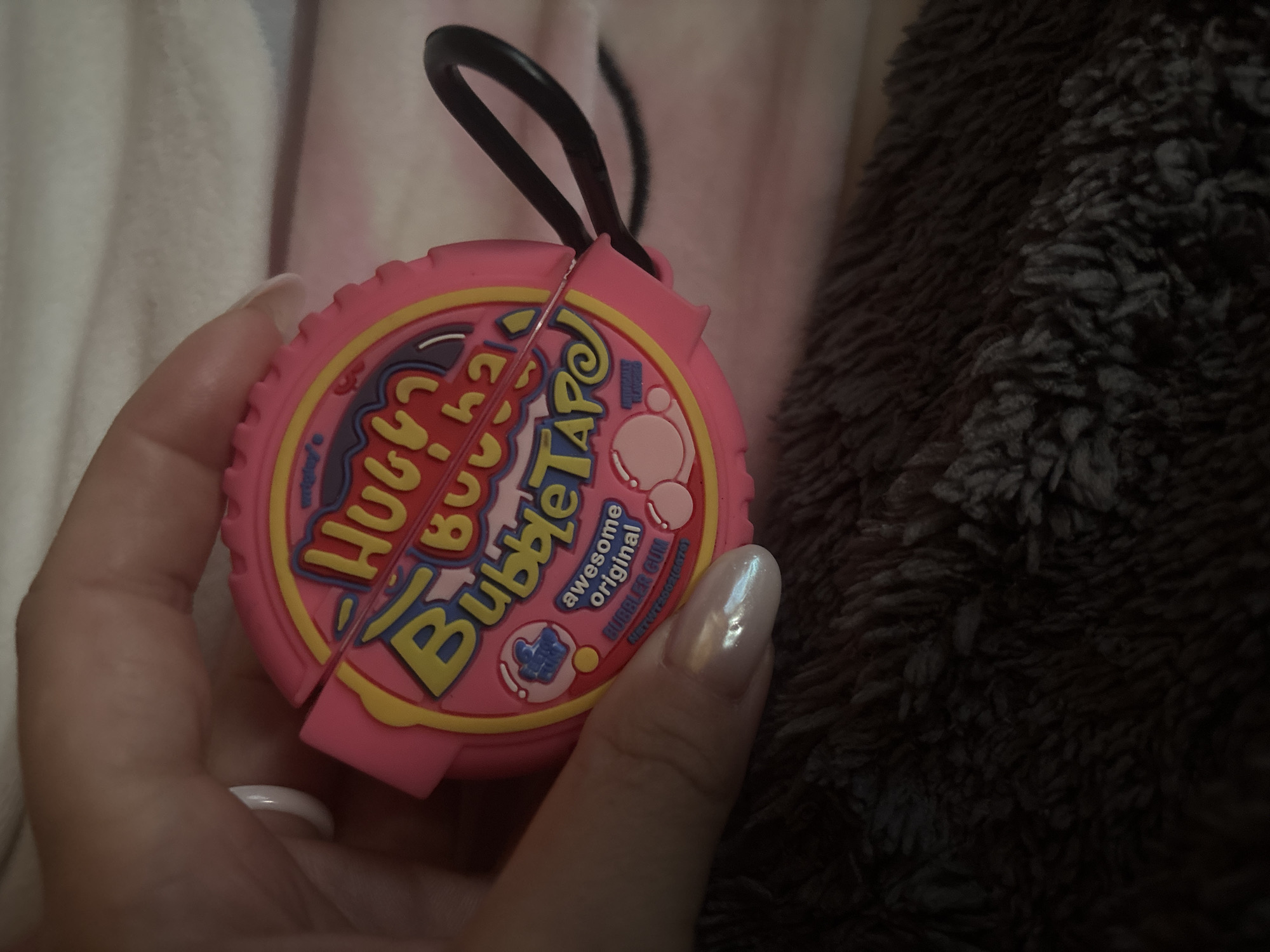 A person is holding a pink, circular object that resembles a roll of bubble gum. The object has a yellow and red design with the words "HUGE BUBBLE TAPE" and "awesome original BUBBLE GUM" printed on it. There is a black loop attached to the side of the object. The person's hand has manicured fingernails. The background is blurred and appears to be a soft, fuzzy surface. There is no indication of Nadezhda, Russia in the image.