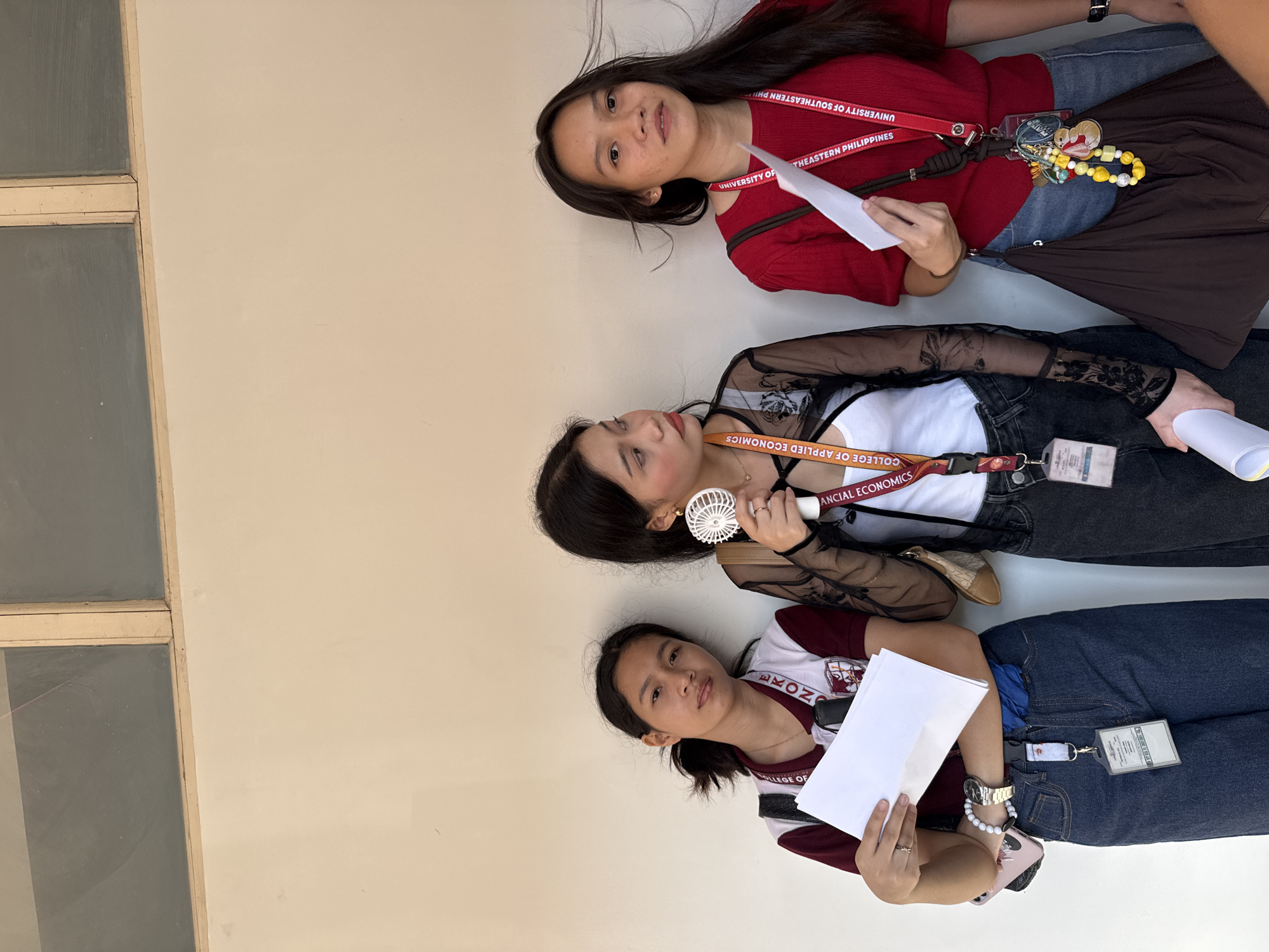 Three young women, presumably students, are positioned on a neutral-colored surface, likely a floor or ground area. They are wearing lanyards that indicate they are associated with the University of Southeastern Philippines. The lanyards are marked with "UNIVERSITY OF SOUTHEASTERN PHILIPPINES" and "COLLEGE OF APPLIED ECONOMICS." They are wearing jeans and casual tops. One woman is holding a white portable fan, and the other two are holding papers. The location is Davao City, Philippines.