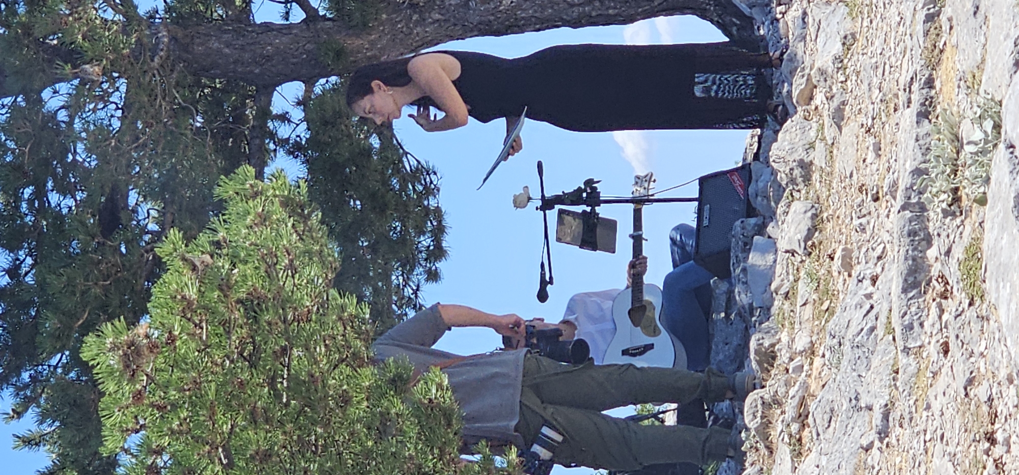 This image depicts an outdoor event taking place on a rocky, arid terrain with sparse vegetation under a clear blue sky. Dominating the scene are large trees with dense green foliage providing shade. In the foreground, on the left, a person is partially visible, wearing a grey t-shirt and olive-green pants, with a camera bag attached to their belt and holding a camera. To the right, a woman in a long black dress stands holding a document, her hand near her face. In the middle ground, a man in a white shirt and blue jeans is seated, playing a white acoustic guitar. A microphone stand with a small decorative item on top is positioned between the guitarist and the woman. A black amplifier with a "BOSS" logo is visible near the guitarist's feet. The overall impression is of a small, informal performance or ceremony in a natural, somewhat rugged environment. The location is identified as being in Općina Bol, Croatia, which is known for its beautiful rocky coastlines and Mediterranean vegetation.