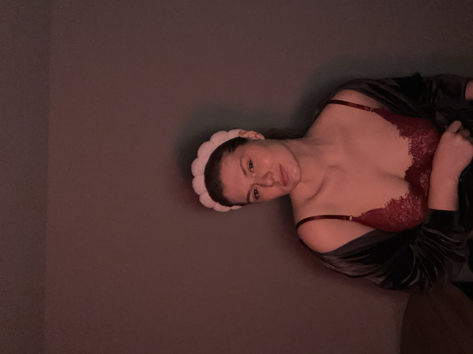 A woman with fair skin and a white puffy headband reclines against a gray wall. She is wearing a burgundy lace bra with thin straps and a dark, velvety robe draped over her arm. The image is dimly lit with a reddish tint. Her head is tilted, and she looks toward the camera with a slight smile.