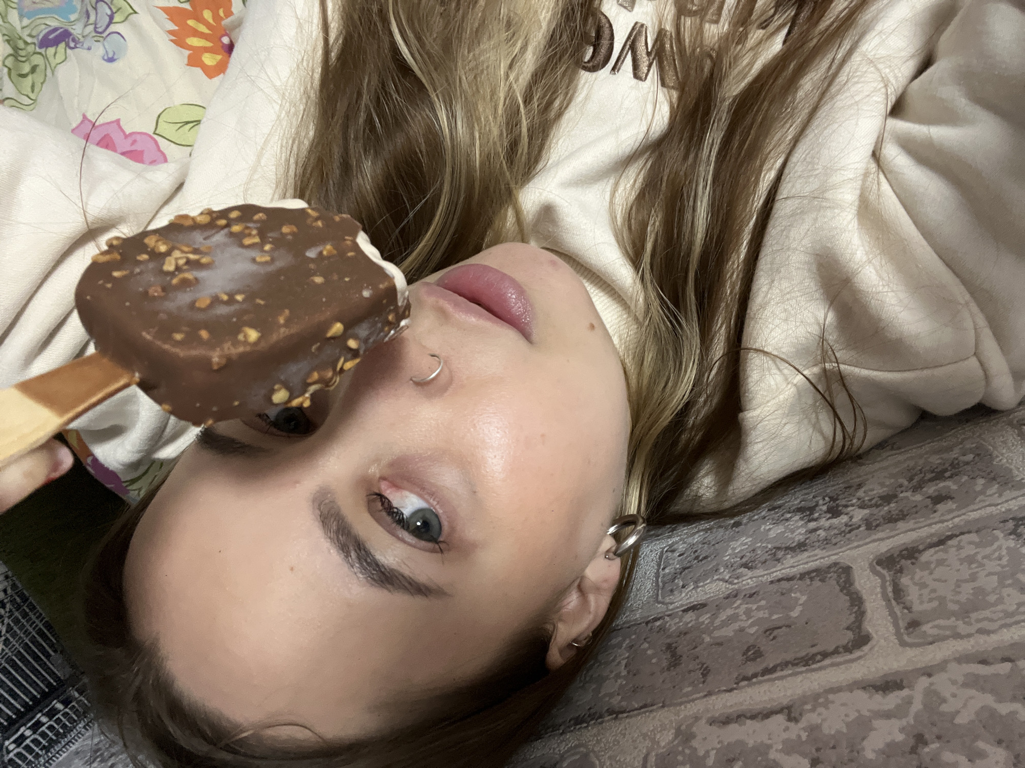 A young woman is depicted in a close-up, partial portrait, lying down indoors in Novopokrovskaya, Russia. Her head is tilted, and her face is viewed from a slightly upward angle, showing her left eye, part of her nose, and her lips. She has long, light brown hair, and a small silver hoop piercing in her nose, along with a large silver ear cuff on her left ear. Her lips are a soft pink.
She is holding a rectangular chocolate-covered ice cream bar with visible chopped nuts, positioned near her face. The ice cream is on a wooden stick. She is wearing a light-colored (beige or off-white) hooded top, with a partial dark brown word, resembling "OM", visible on the fabric.
The background features two distinct patterns: behind her upper head is a light-colored fabric with a vibrant floral design in pink, orange, and green. To her side and below her, there is a grey surface with a repeating brick-like pattern. The lighting suggests an indoor setting.