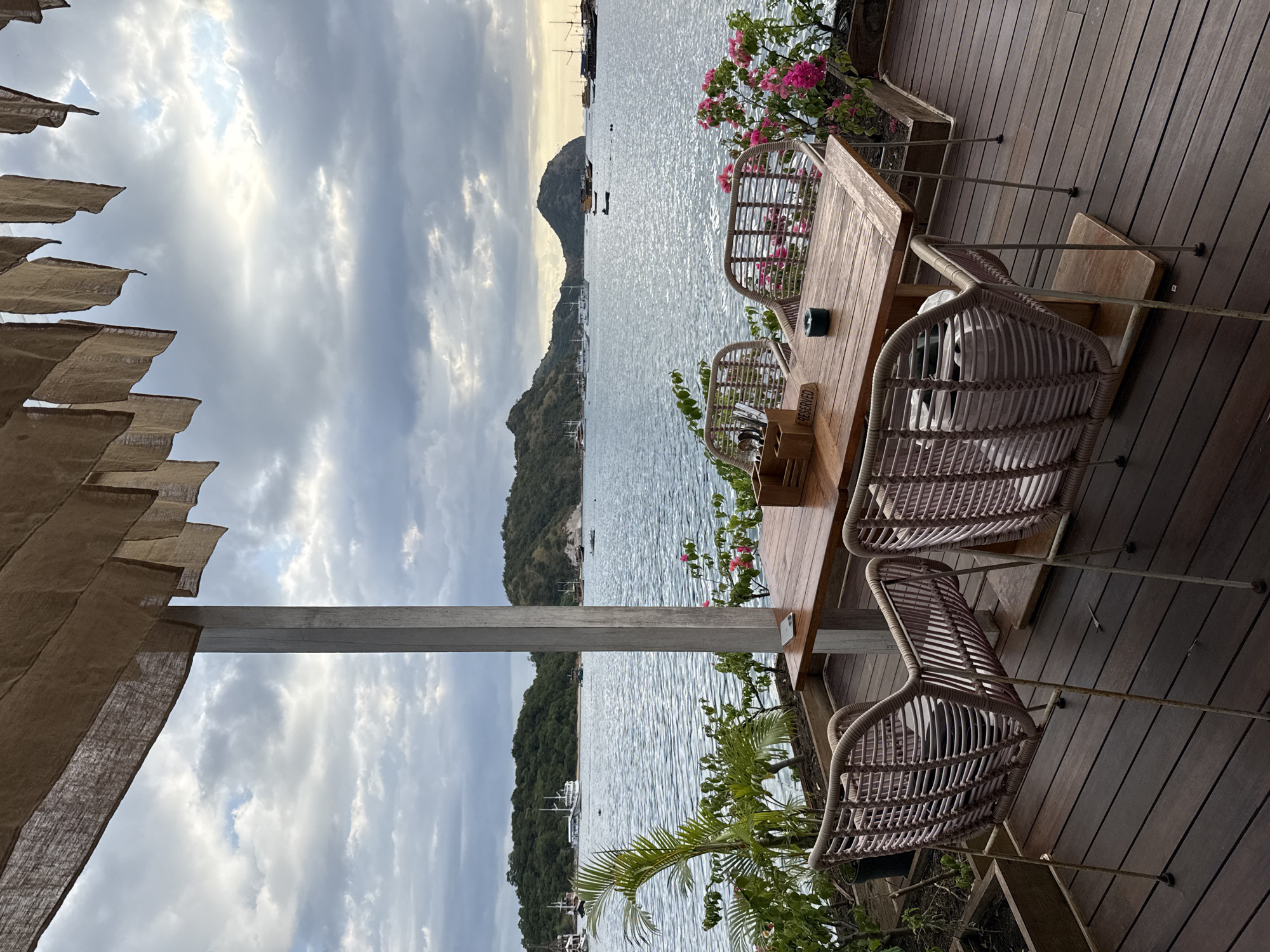 The image shows an outdoor seating area overlooking a bay in Labuan Bajo, Indonesia. A wooden table and four wicker chairs are arranged on a wooden deck. Plants with pink flowers decorate the table and deck area. In the background, the bay is visible, with islands and boats dotting the water. The sky is overcast with grey clouds. A beige fabric awning is visible in the foreground on the left. A metal support beam bisects the view of the bay. A “Reserved” sign is on the table.