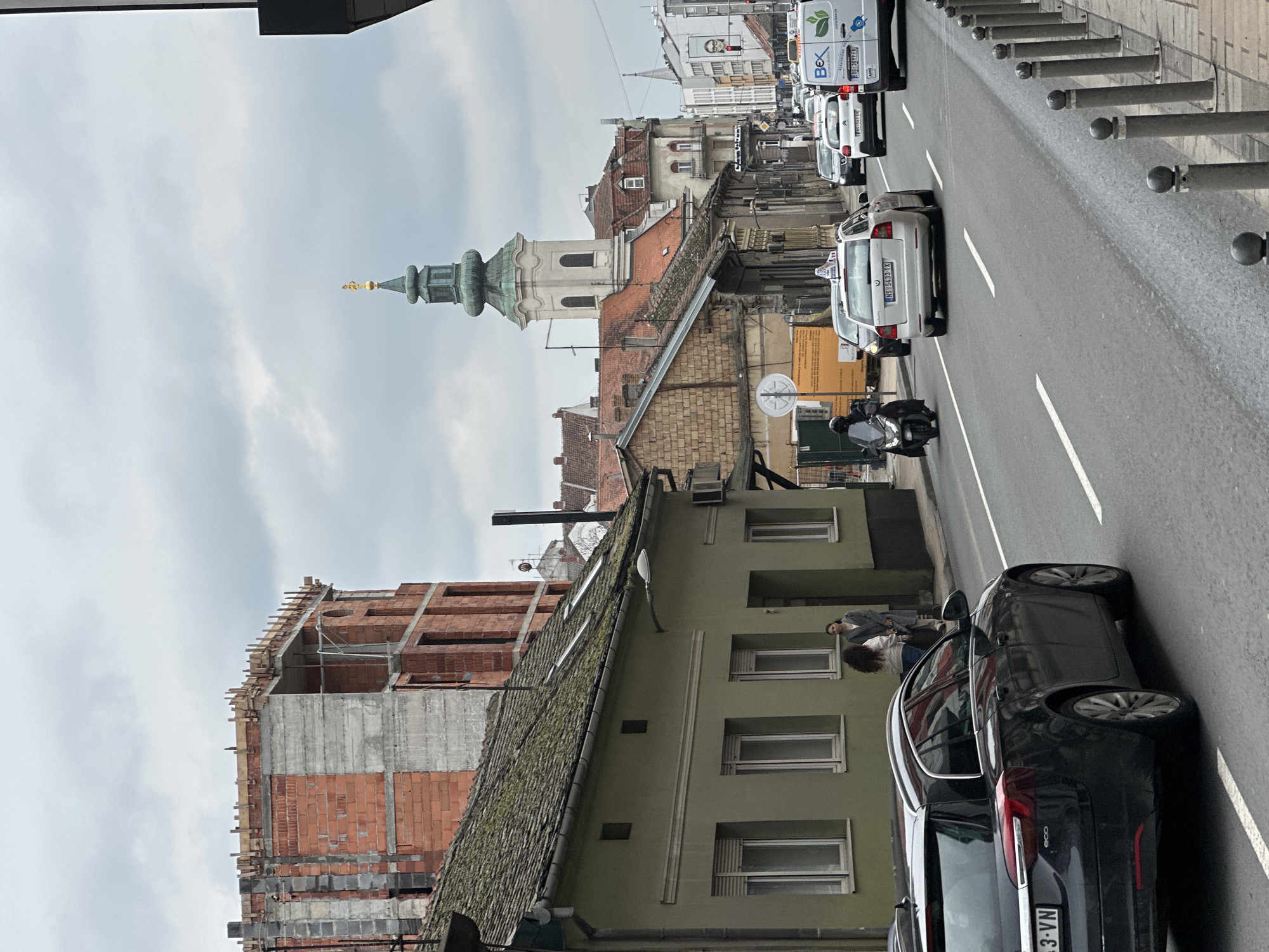 This image shows a street in Novi Sad, Serbia, featuring a mix of old and new architecture. A church tower rises in the distance among red-roofed buildings. In the foreground, a building under construction with exposed brick and scaffolding stands next to an older, green building with multiple windows.
A dark-colored car is parked on the right side of the street with license plate number "NS 223-VN". Two people are standing beside the car, one wearing a light-colored jacket and jeans with curly dark hair, and another wearing a grey suit.
The street has marked lanes and a sidewalk with metal bollards. Several cars and vans are moving along the street. The sky is overcast.