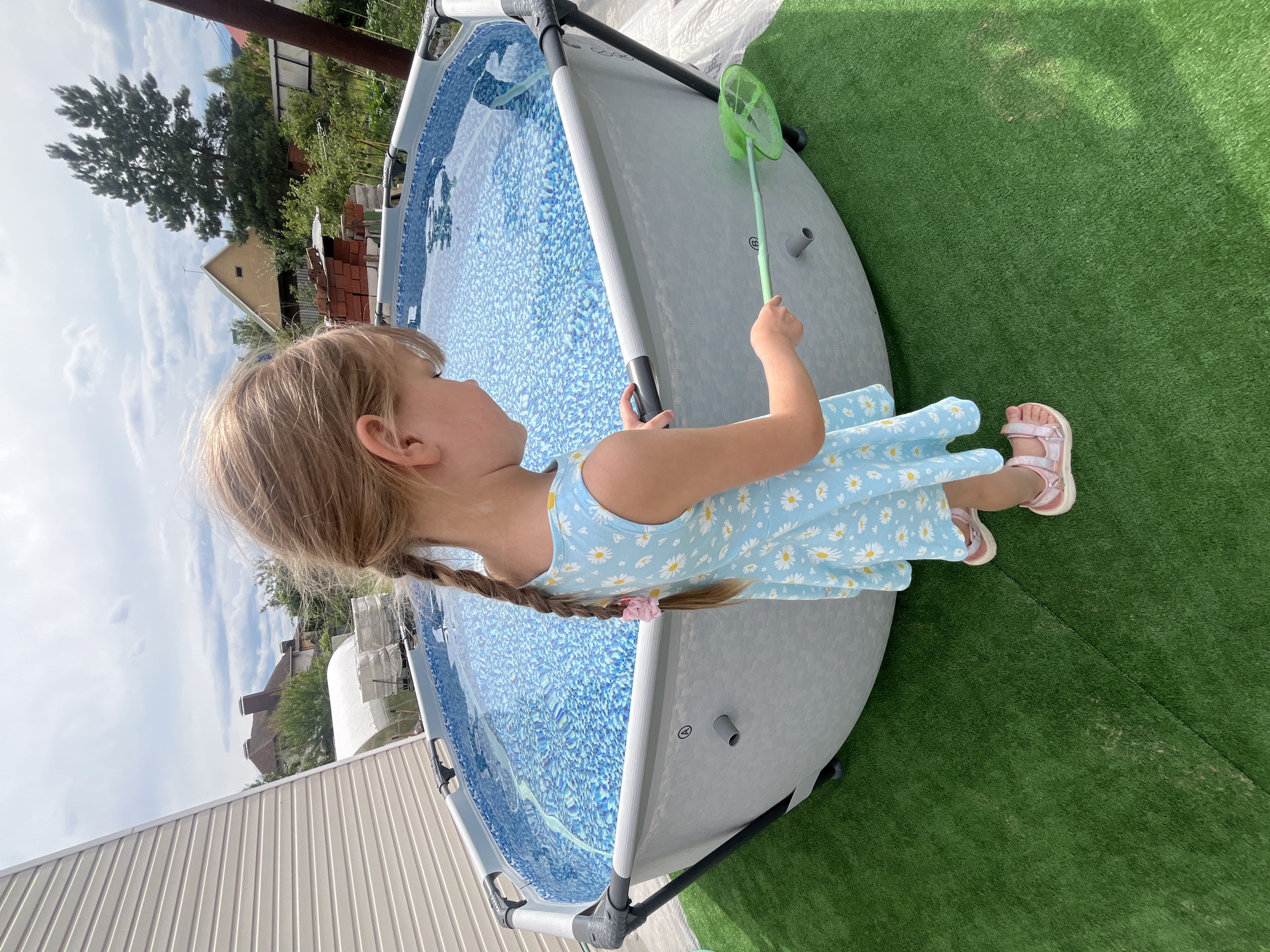 A young Caucasian girl with blonde hair braided into two pigtails, wearing a light blue dress with a daisy pattern and pink sandals, stands on artificial green turf next to a circular, above-ground swimming pool. She is facing away from the camera, looking into the pool, and holding a green net on a long handle. The pool is filled with blue water with a white patterned liner. The background shows a residential area with houses, trees, and a cloudy sky. This scene is likely in a backyard in the city of Voronezh, Russia, given the architectural style of the houses and vegetation.