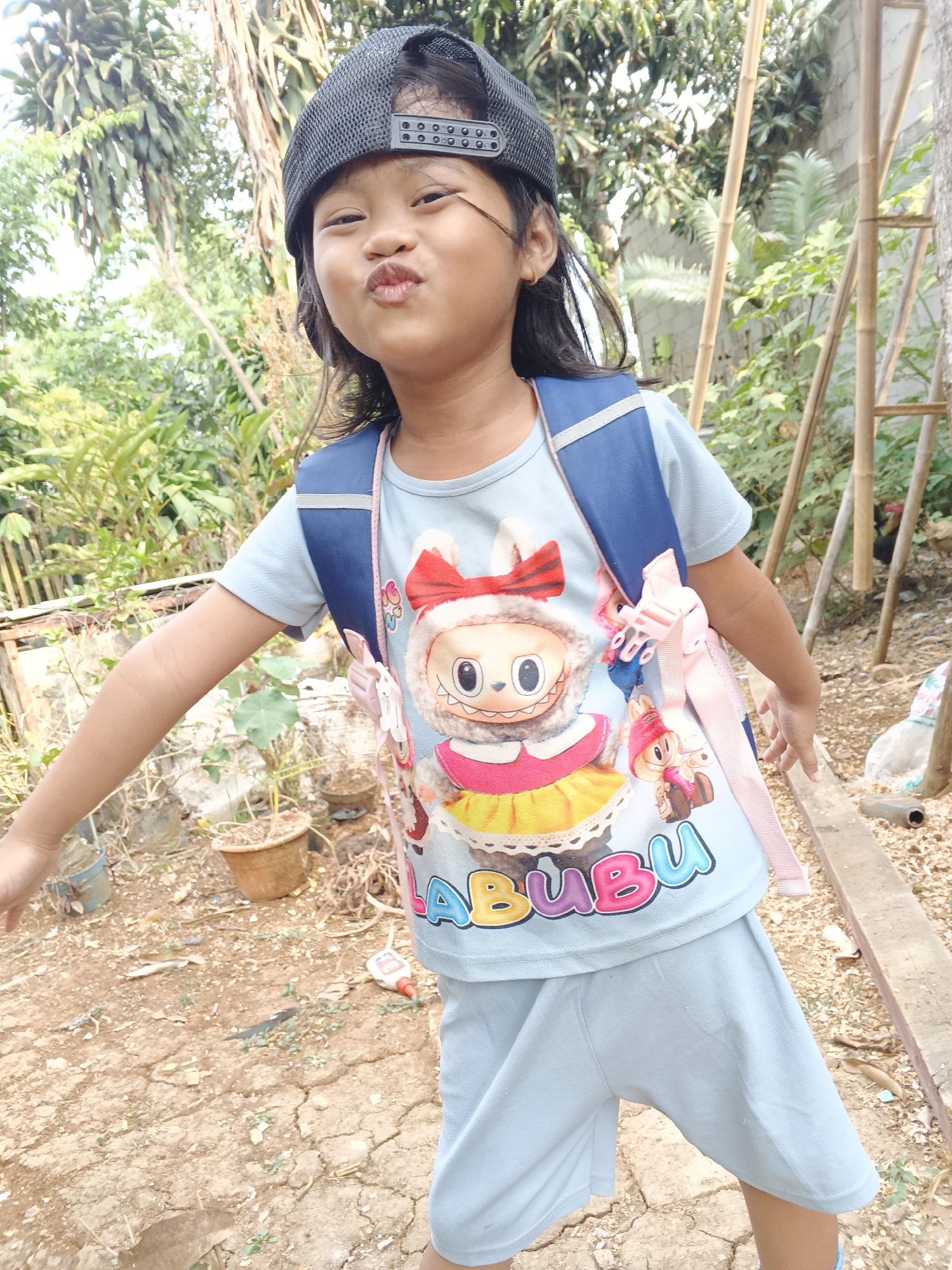 A young East Asian female, wearing a light blue t-shirt with a cartoon character and "BUBU" text, light blue shorts, and a black baseball cap, stands in an outdoor environment with dry, cracked earth. She is facing the camera with her mouth puckered and has a blue backpack with pink straps on her back. The background consists of vegetation, bamboo poles, a brick wall, and various potted plants. The scene appears to be in a backyard or outdoor area within the city of Warungkiara, Indonesia, given the style of the vegetation and building materials.