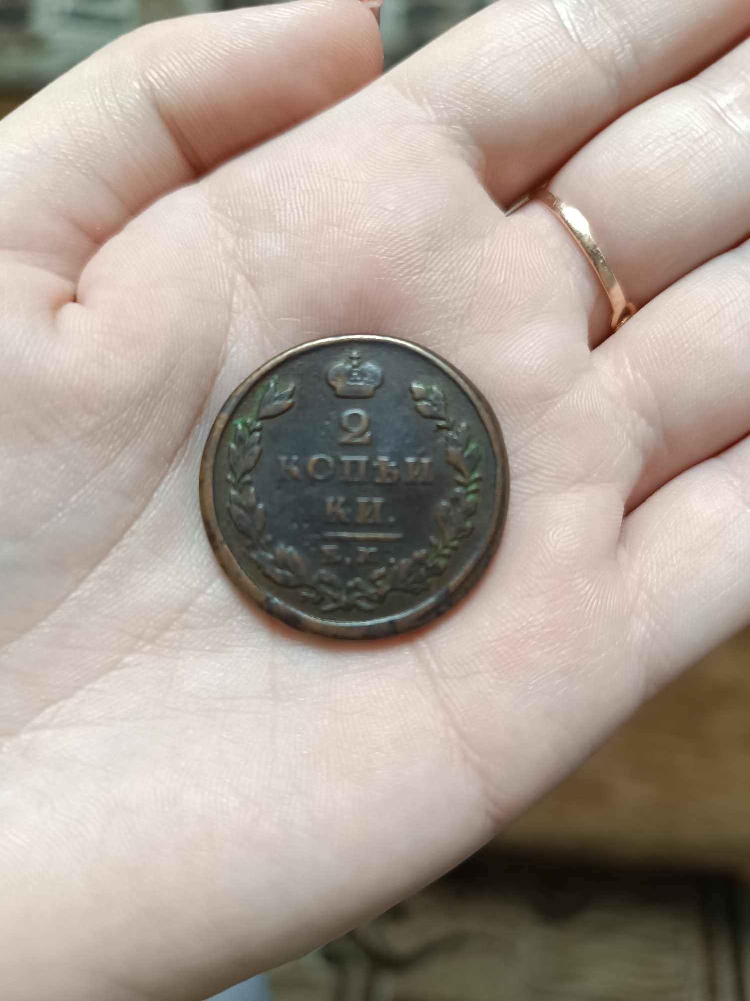 A close-up shot shows a palm holding a worn, circular coin. The coin features the inscription "2 копѣи" (2 Kopeks) in Cyrillic script, along with a crown at the top and what appear to be laurel branches flanking the text. There's also the Cyrillic inscription "К.И." and "Е.М." below. The coin appears to be of historical significance, potentially from Imperial Russia. The hand is light-skinned, with a simple gold ring visible on one finger. The background is blurred and out of focus, suggesting a close-up shot. The city of Chaikovsky, Russia is not identifiable from this image.